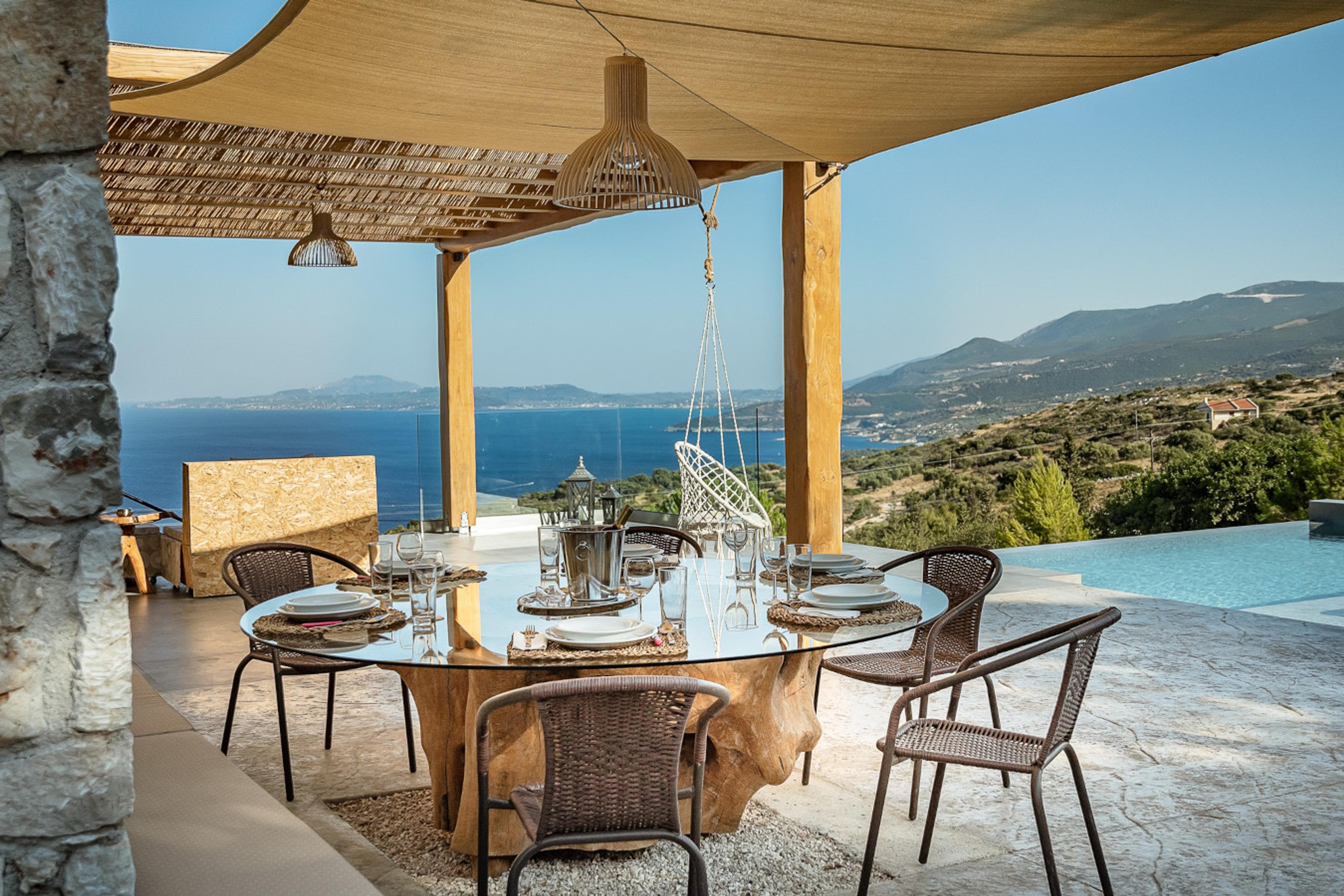 A round wooden dining table laid with plates, wine glasses and a wine bucket under a pergola, framed by the sea and hills beyond.