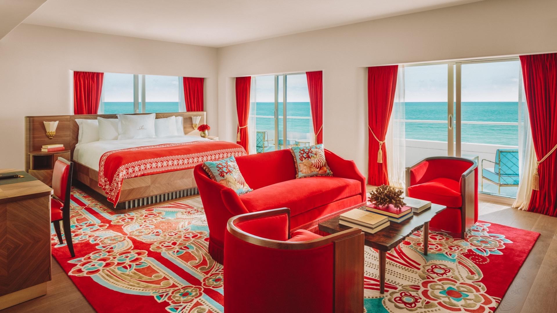 A bedroom with red velvet curtains, a red and white throw on a wooden bed, a red sofa and armchairs on a patterned rug, and the sea visible through the balcony doors.