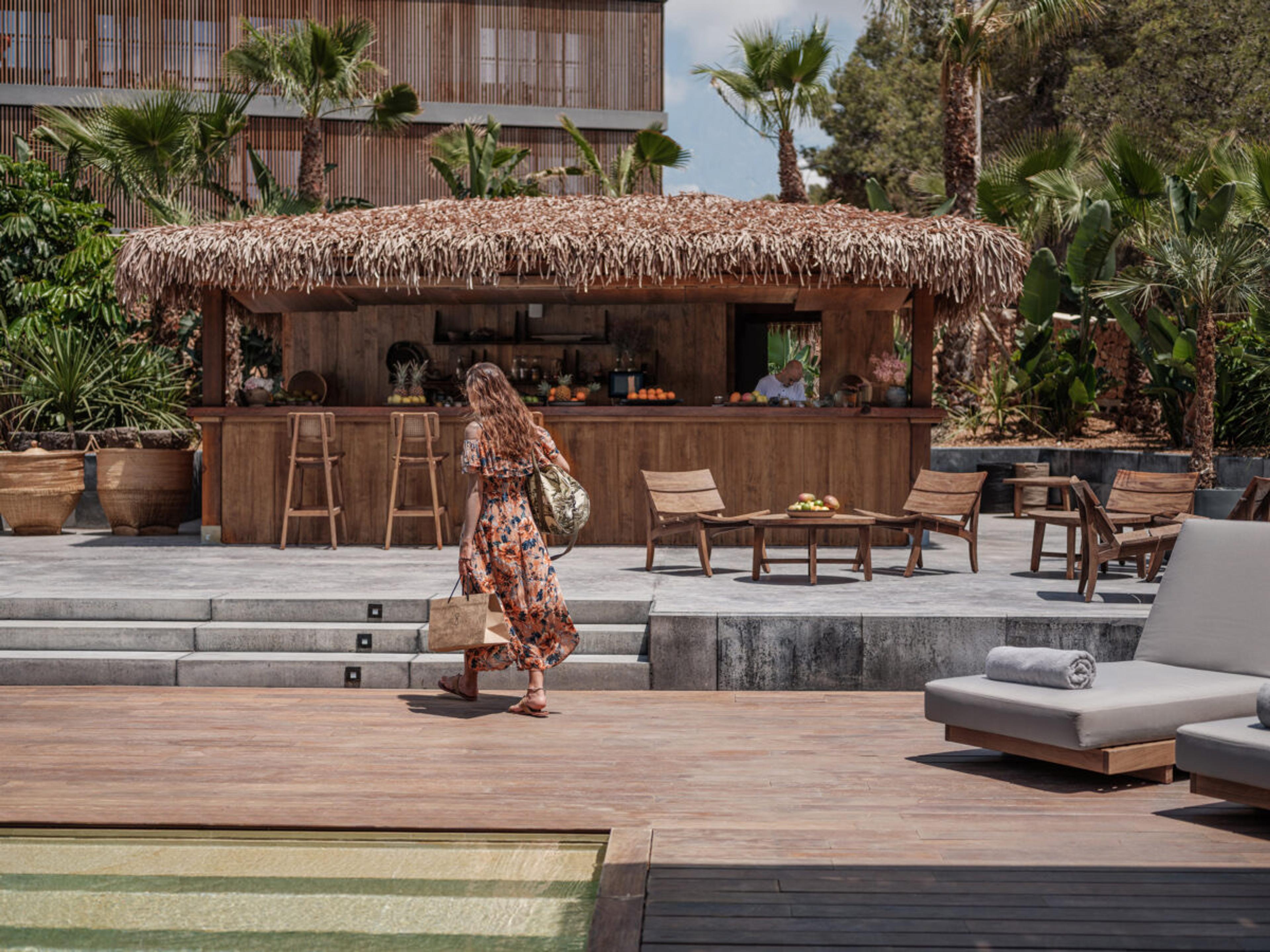 A thatched bar with a timber counter and stools beside a pool deck, with a woman in a floral dress walking away from the camera past a low table and two chairs.