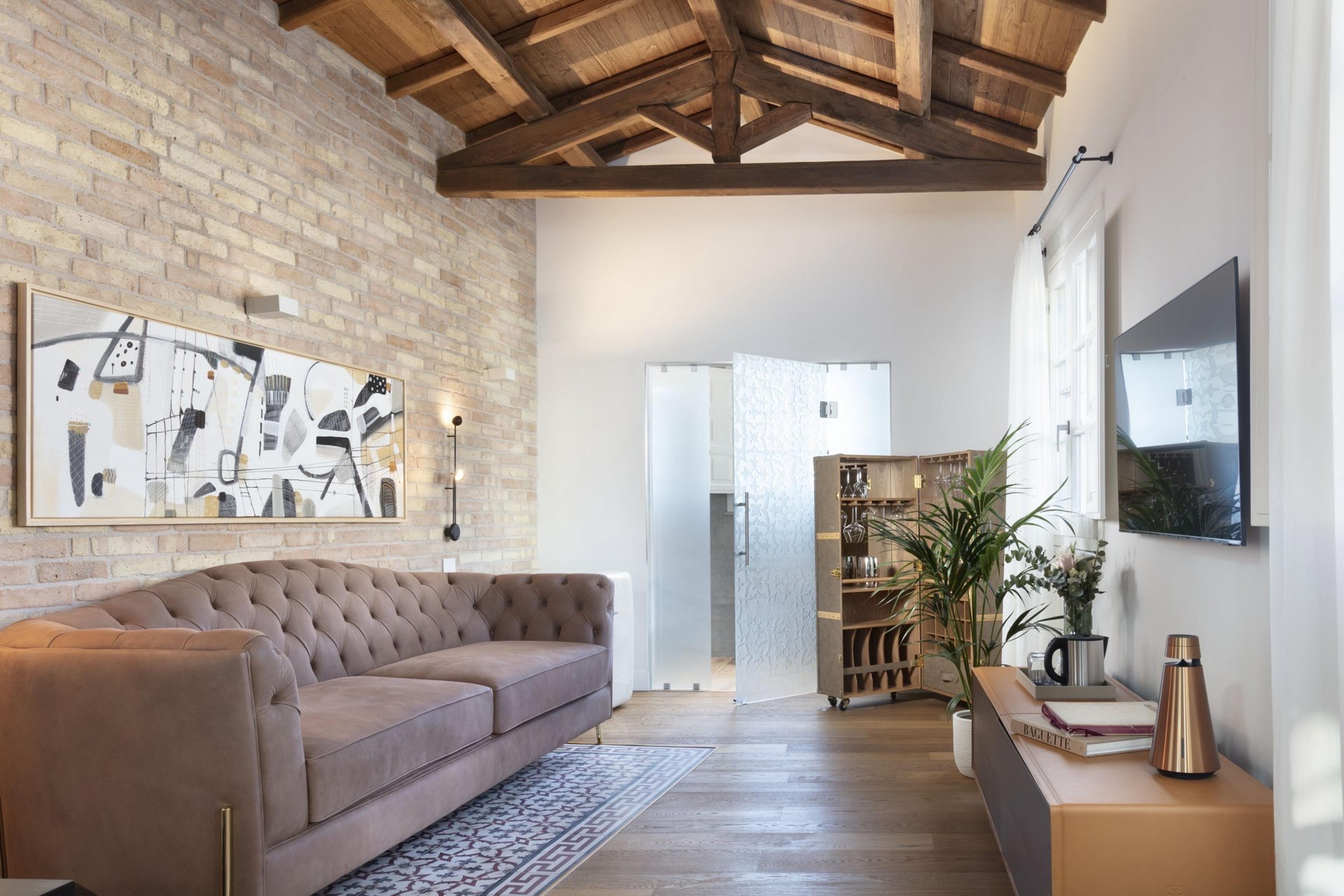 A living room with an exposed brick wall, a tufted leather sofa, an abstract painting, a wall-mounted television, and a wooden drinks trolley by the window.