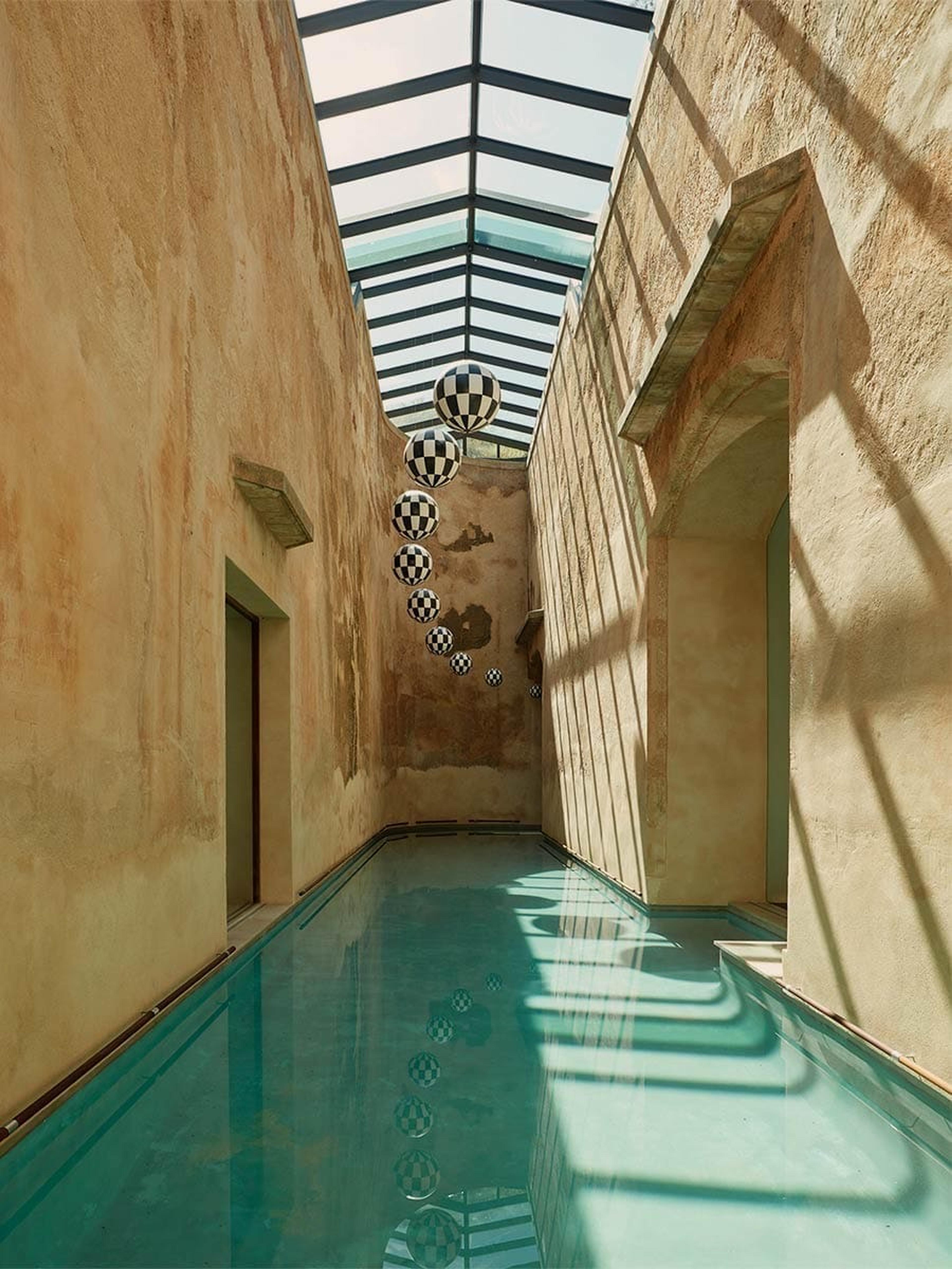 An indoor pool corridor with weathered ochre walls, a glass skylight, and a line of checkered sphere decorations hanging above the water.