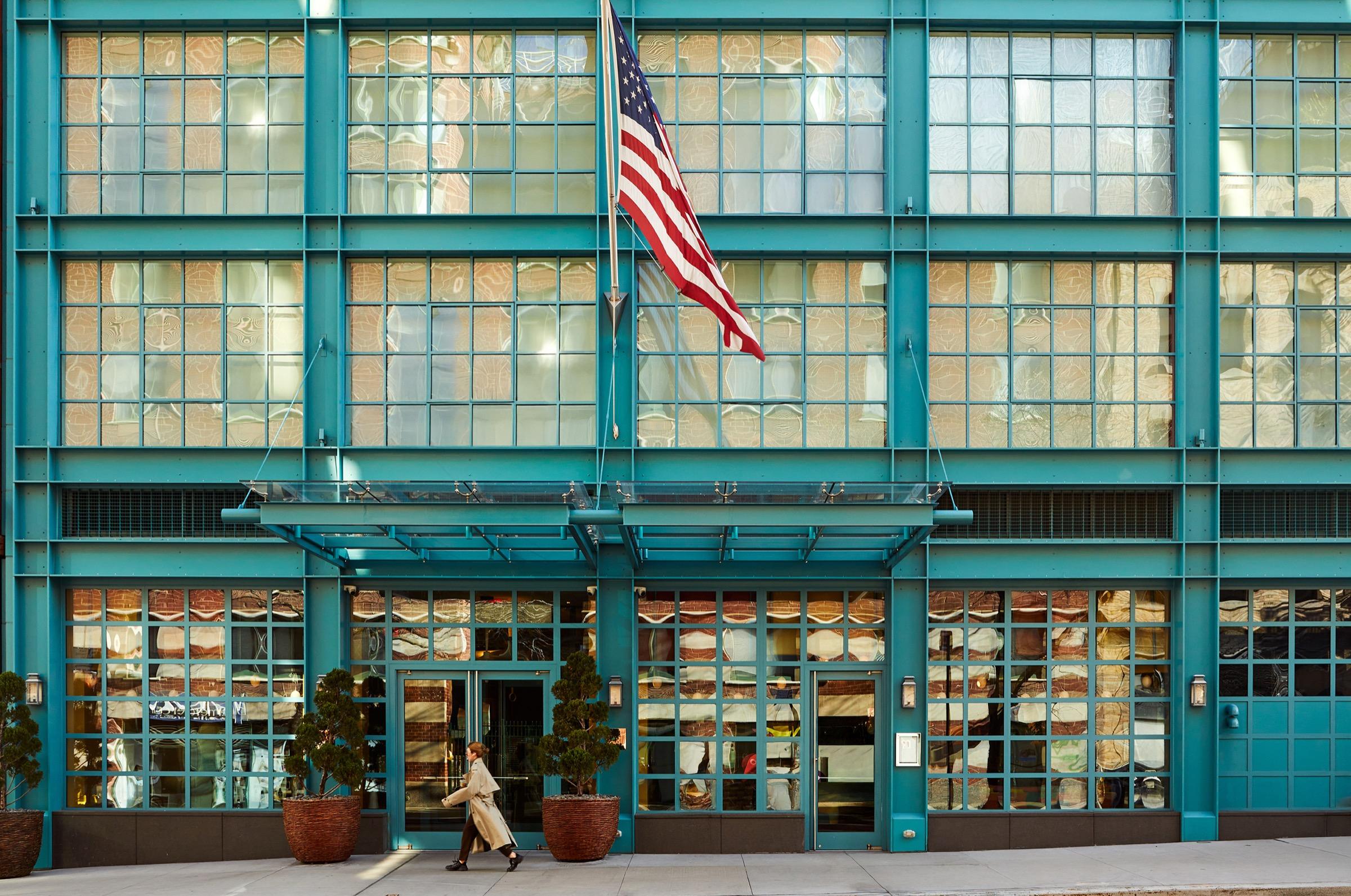 A teal steel-framed hotel facade with floor-to-ceiling industrial windows, an American flag above the entrance, and a woman in a trench coat walking past on the pavement.