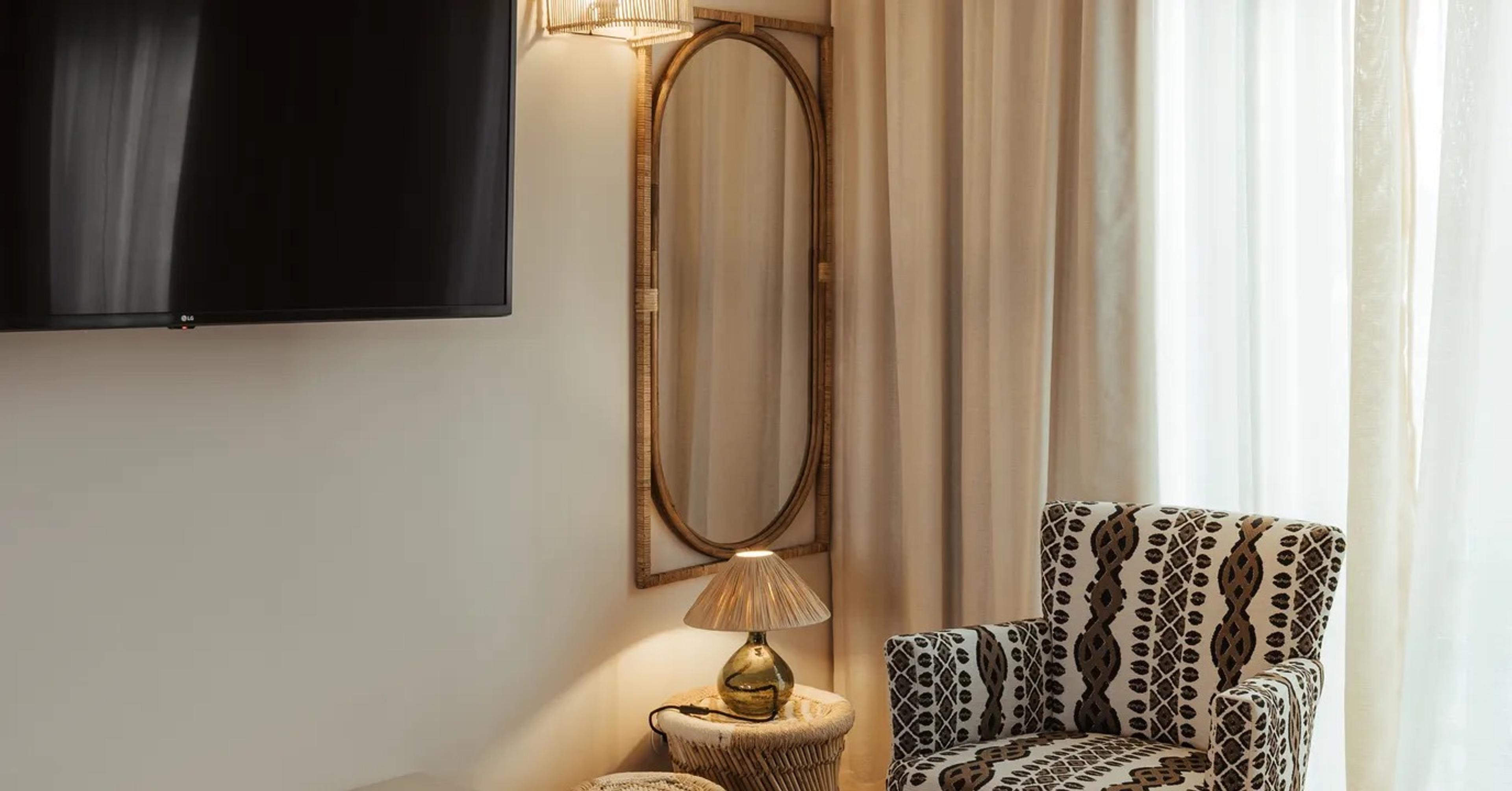 A bedroom corner with a wall-mounted television, an oval rattan-framed mirror, a small table lamp, and a patterned armchair beside sheer curtains.