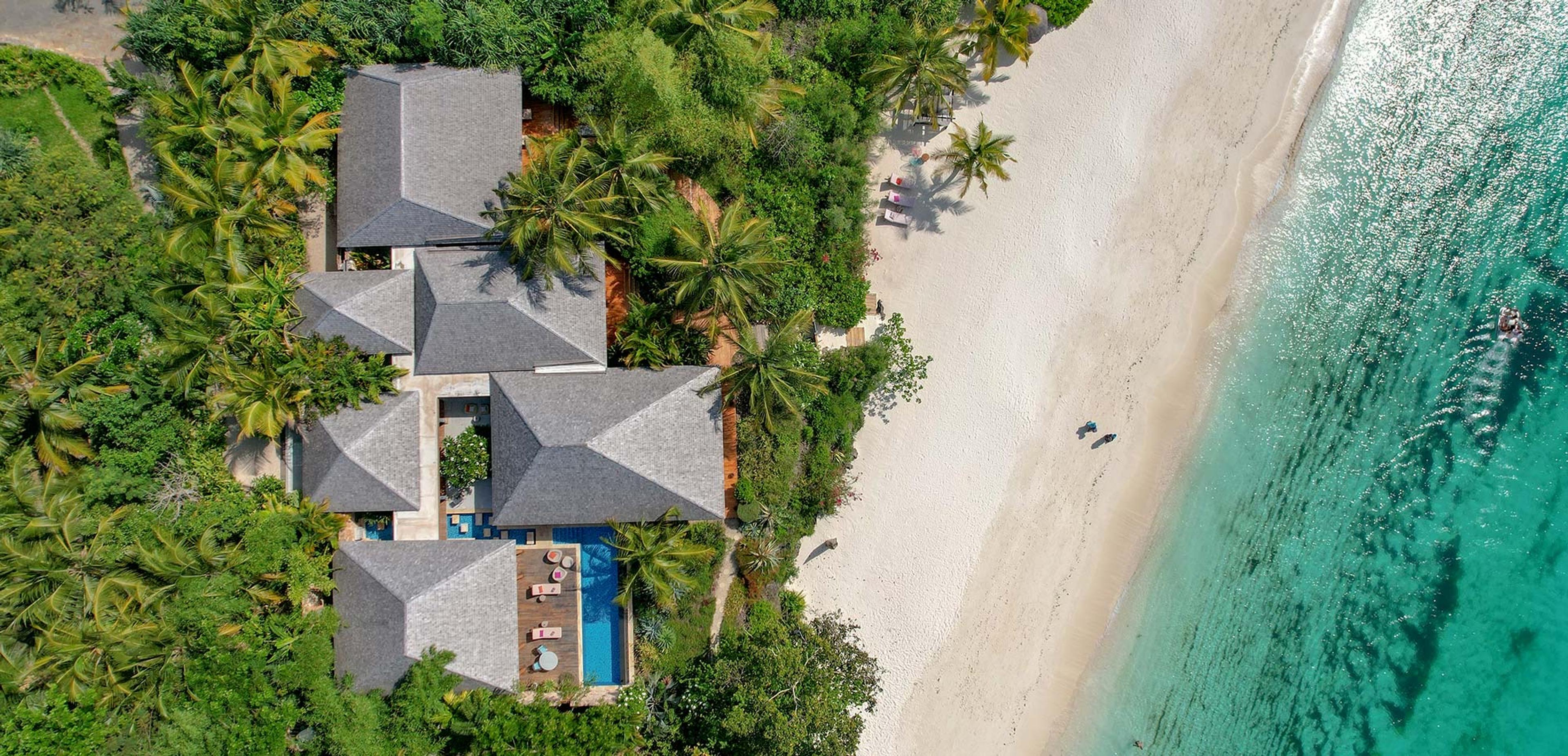 an aerial view of a cluster of thatched-roof villas and a pool among palm trees, right beside a white sand beach and turquoise sea