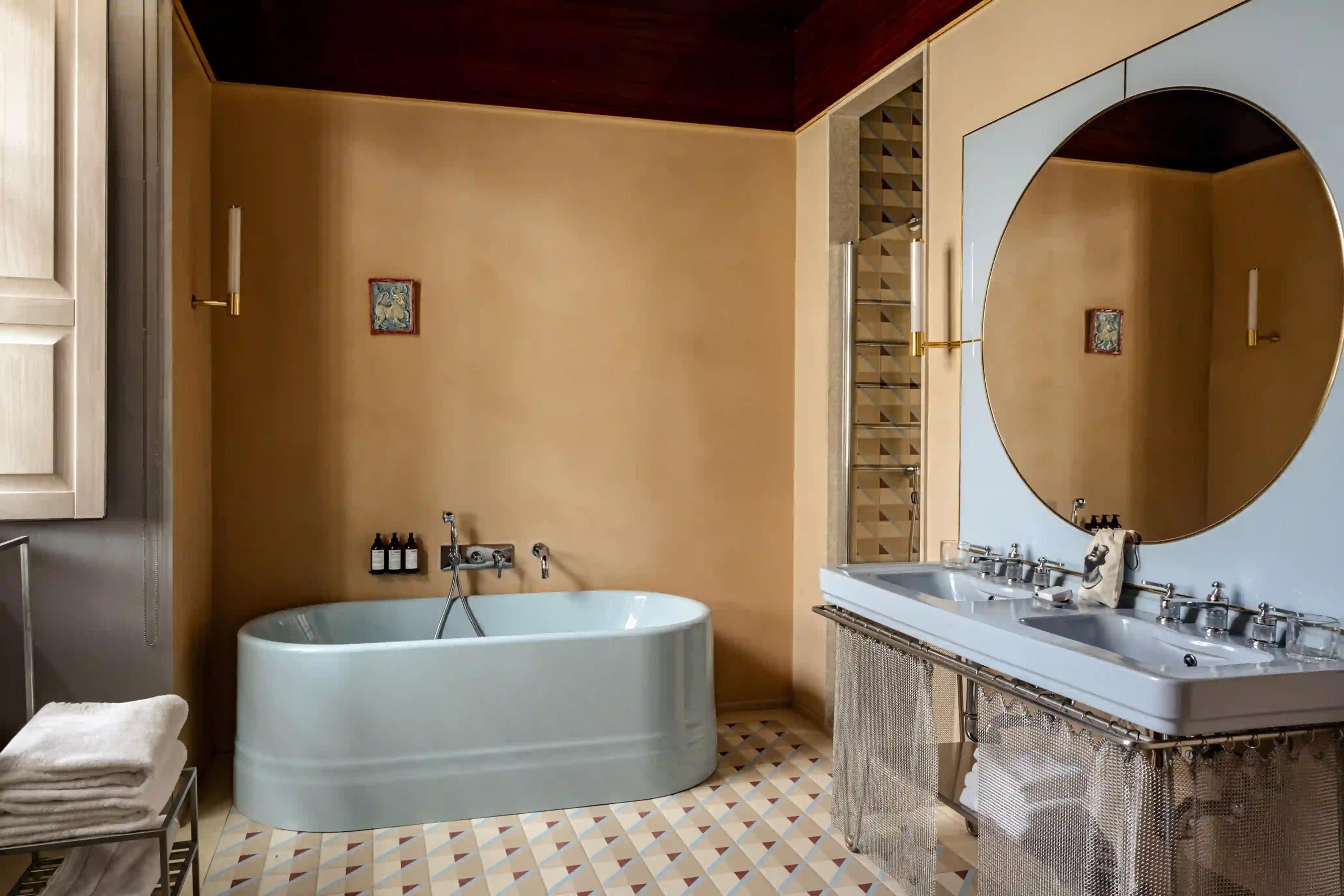 A pale blue freestanding bathtub beside a double sink under a round mirror, on a patterned tiled floor beneath a dark wood ceiling.