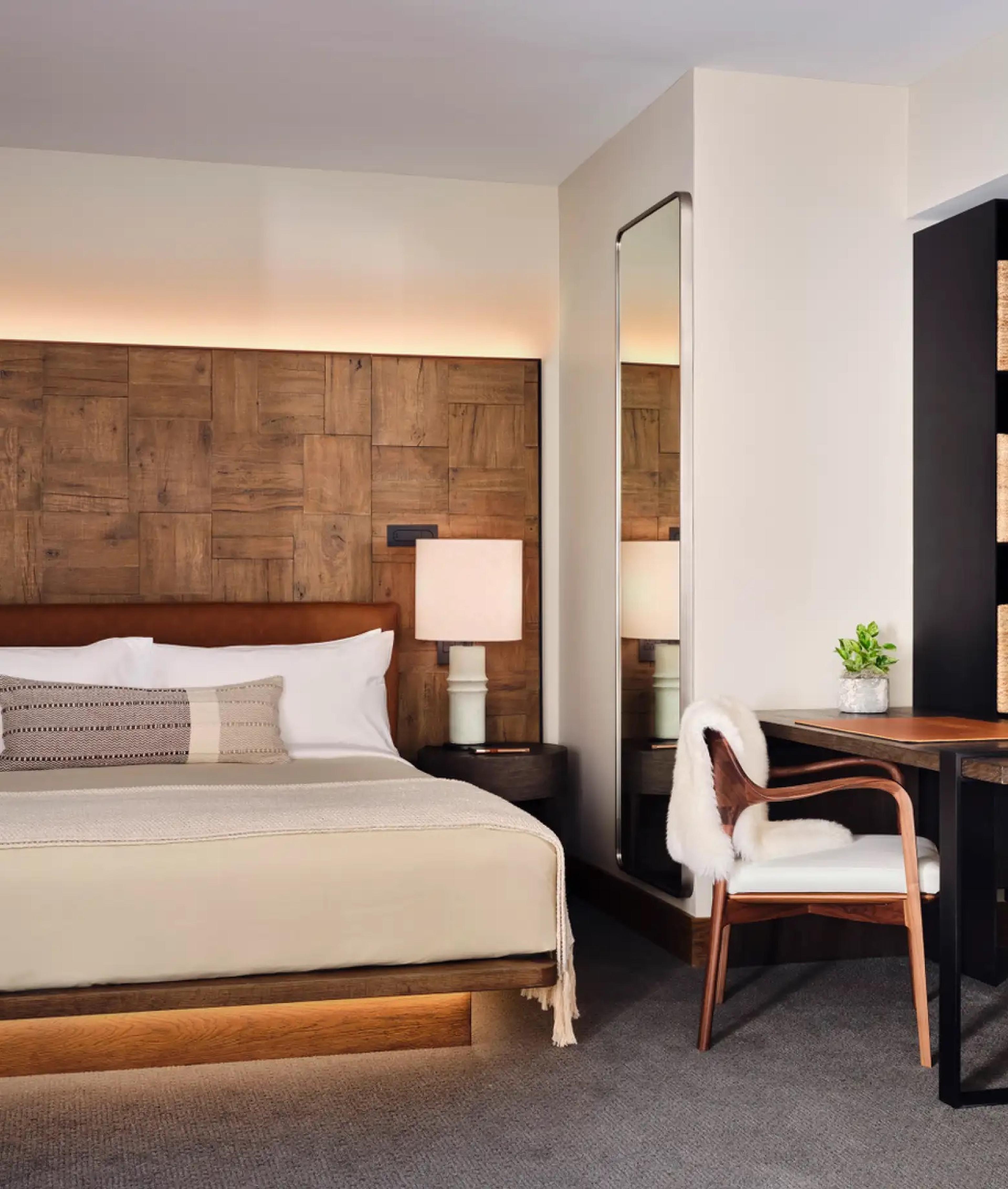 A king bed against a wood-panelled wall, with a table lamp on the bedside table, a tall mirror, and a wooden desk with a white-seated chair.