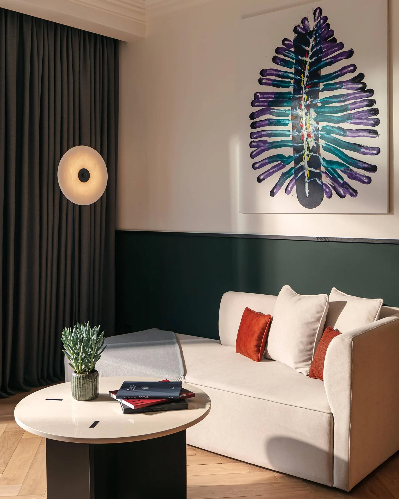 A suite sitting area with a cream sofa, orange and red cushions, a round white coffee table with books, and a large colourful painting above green panelling.