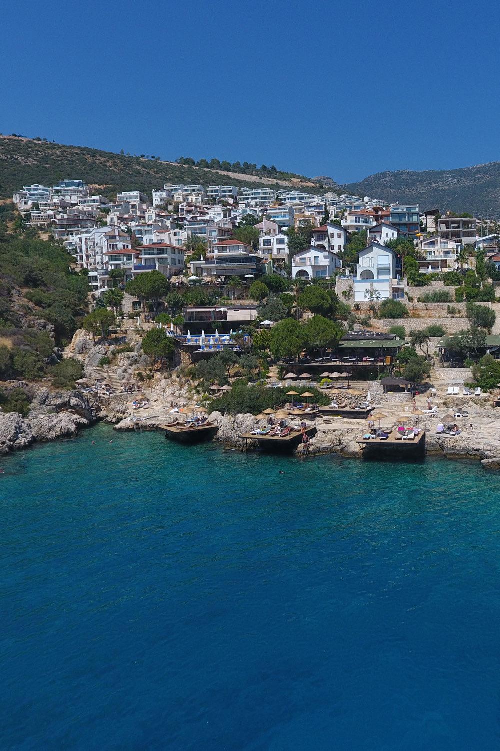 An aerial view of a white hillside town above a bay, with wooden sun platforms and sunbeds built into the rocks at the water's edge.