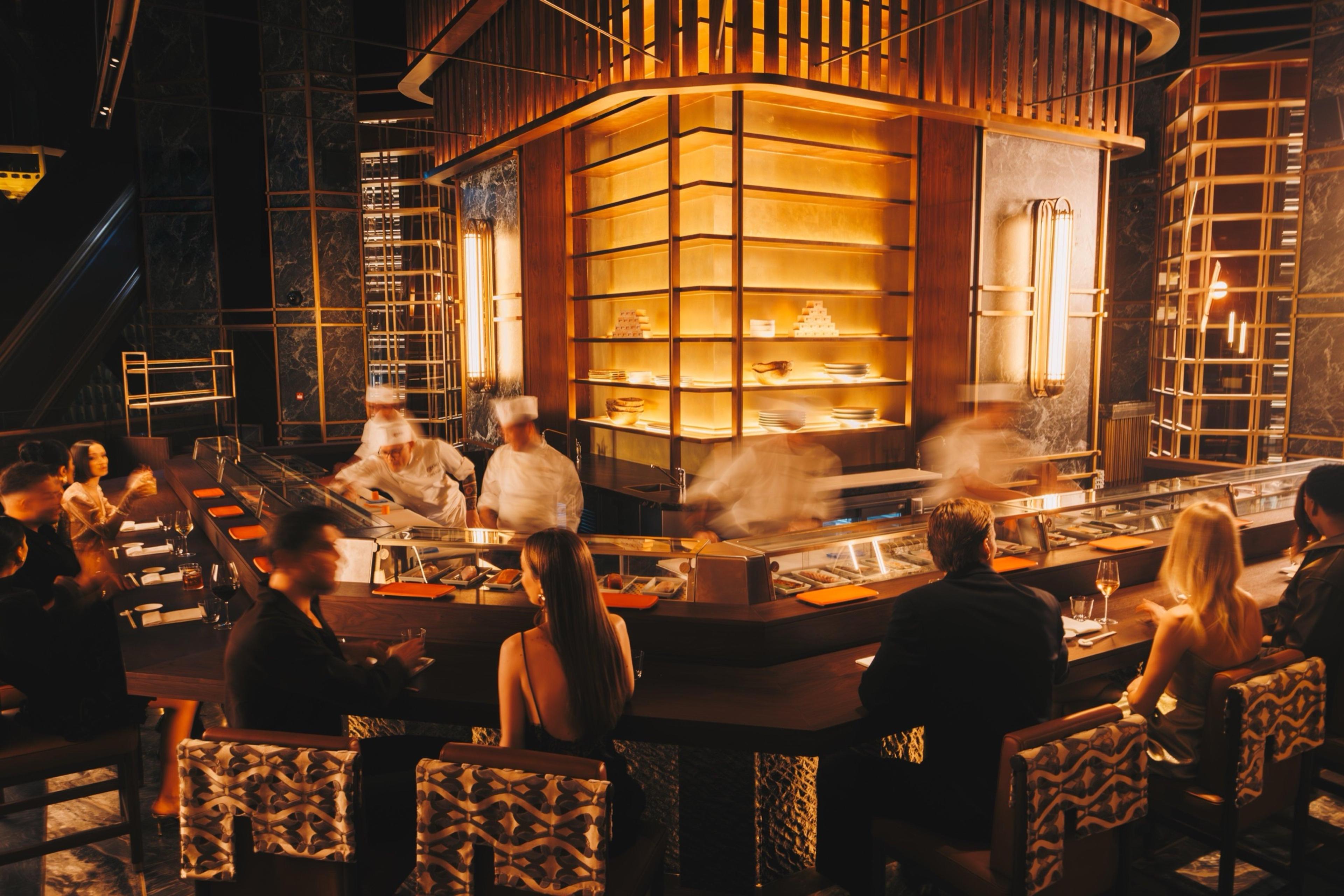 A sushi and robata counter lit in gold, with chefs in white moving behind the bar and guests seated at the counter and surrounding tables.