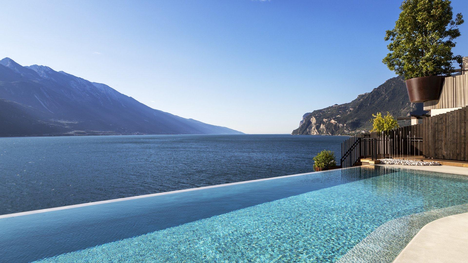 an empty infinity pool against a wide lake and mountain view, with a potted tree at the poolside