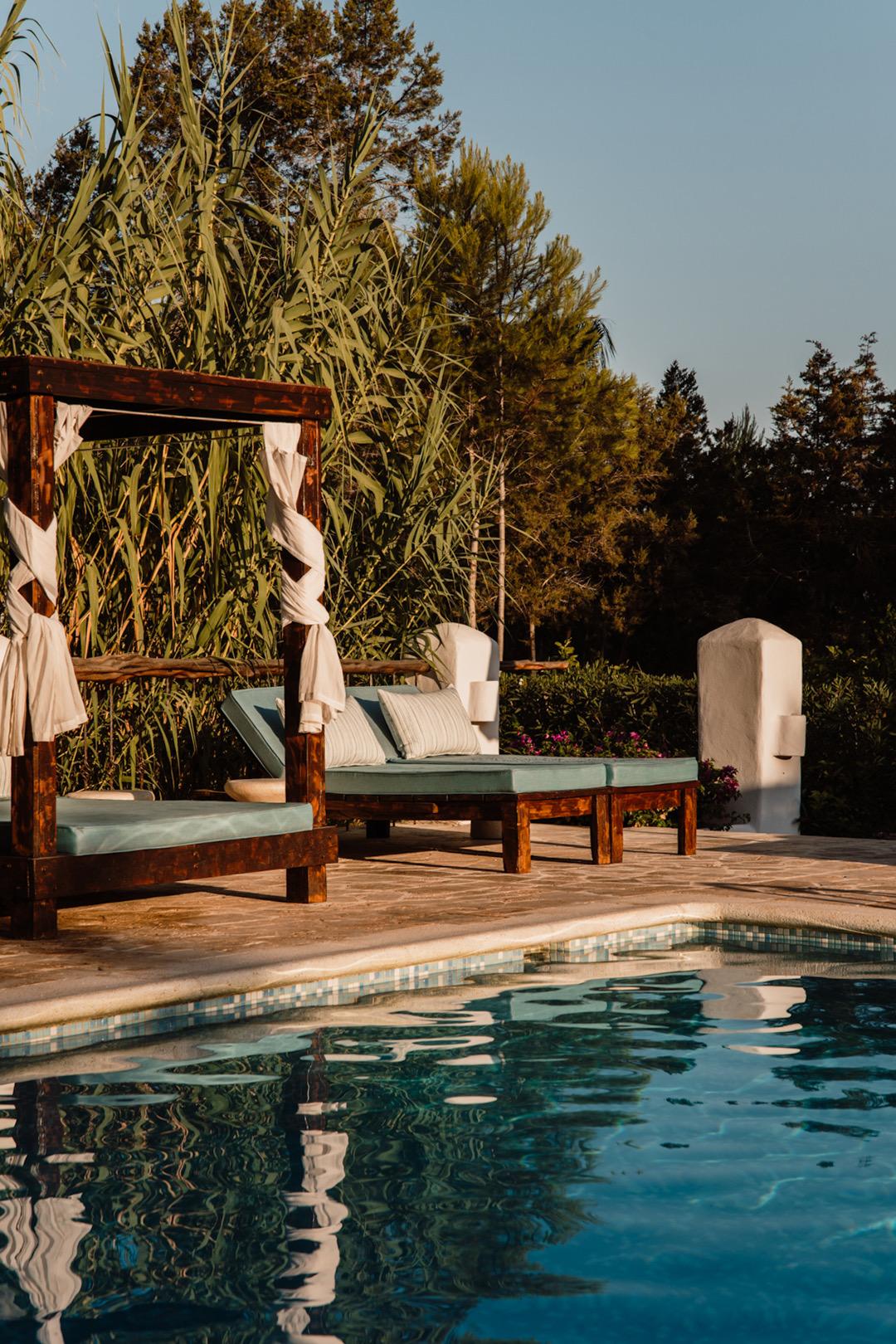 A wooden four-poster daybed and lounger beside a swimming pool, with tall reeds and trees behind, lit by late afternoon sun.
