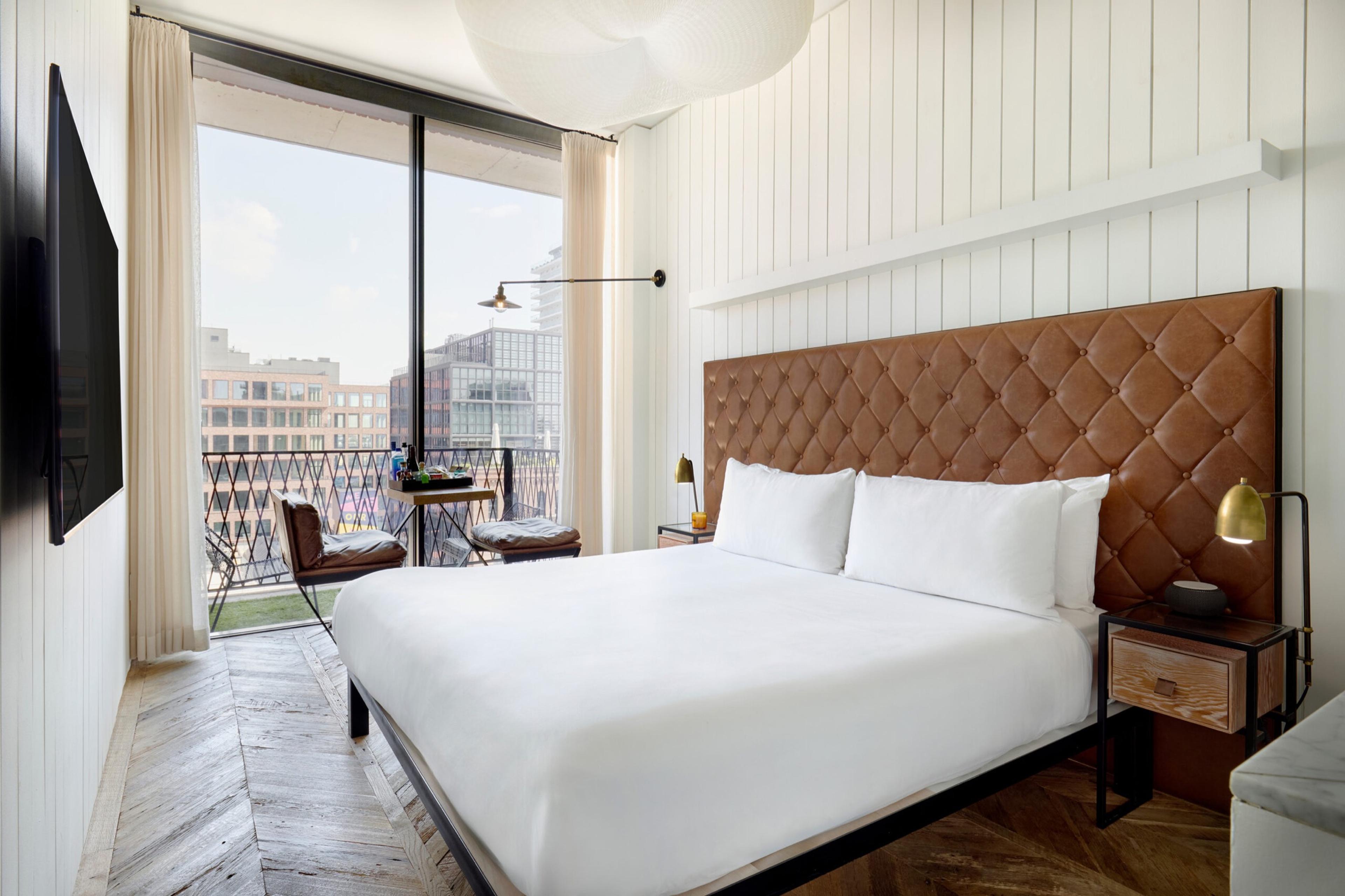 A bedroom with a leather headboard bed, a wall-mounted television, and a glass door open to a balcony overlooking city rooftops.