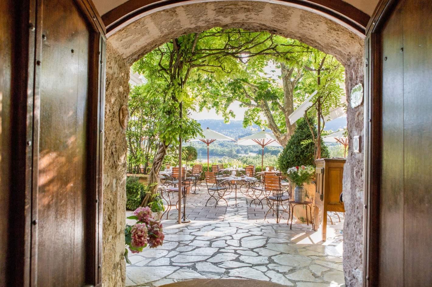 A stone archway framing a wisteria-covered terrace of tables and umbrellas, with wooded hills beyond.