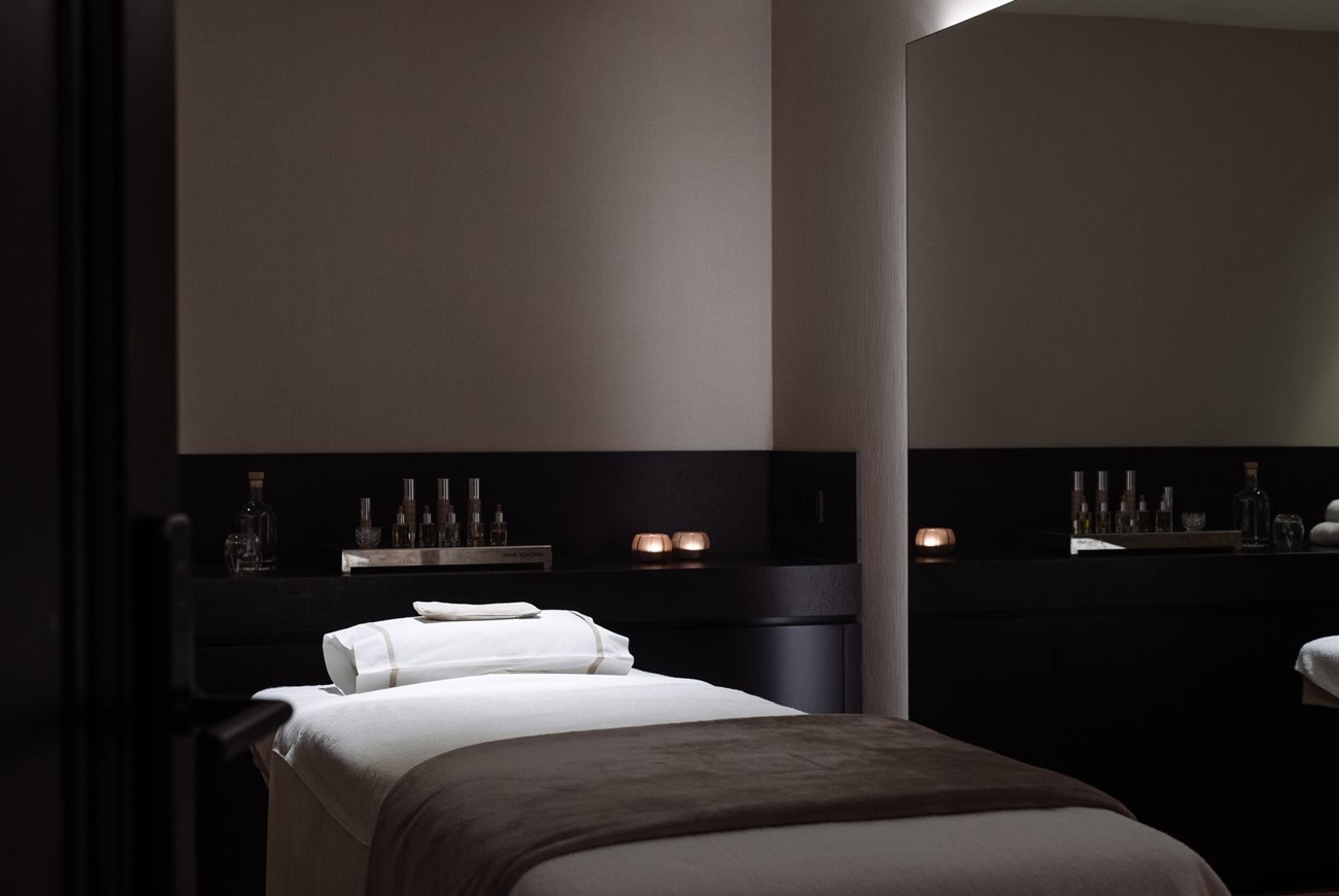 A dark treatment room with a massage table, folded white towels, and bottles of oil lit by candles on a side counter.