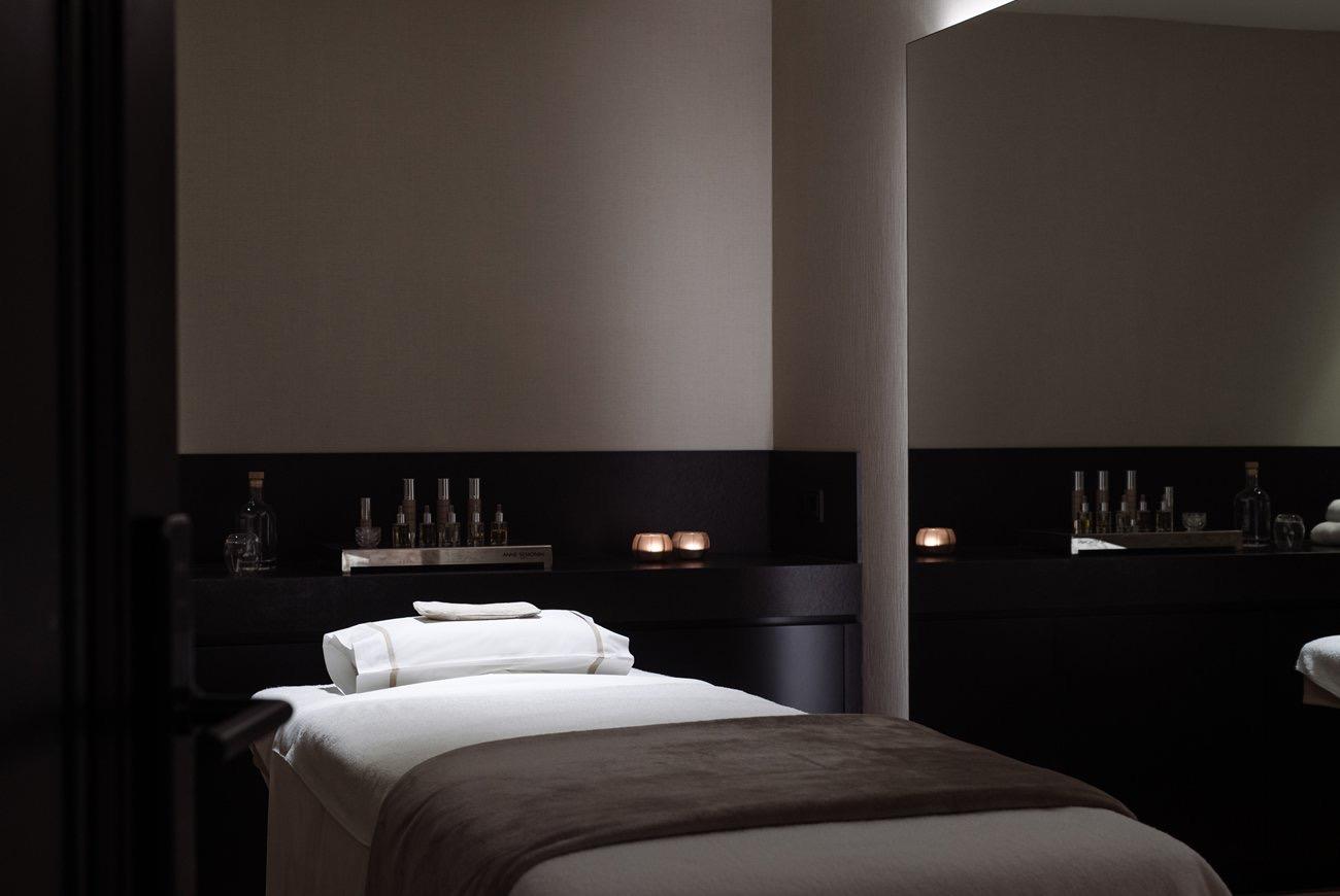 A dark treatment room with a massage table, folded white towels, and bottles of oil lit by candles on a side counter.