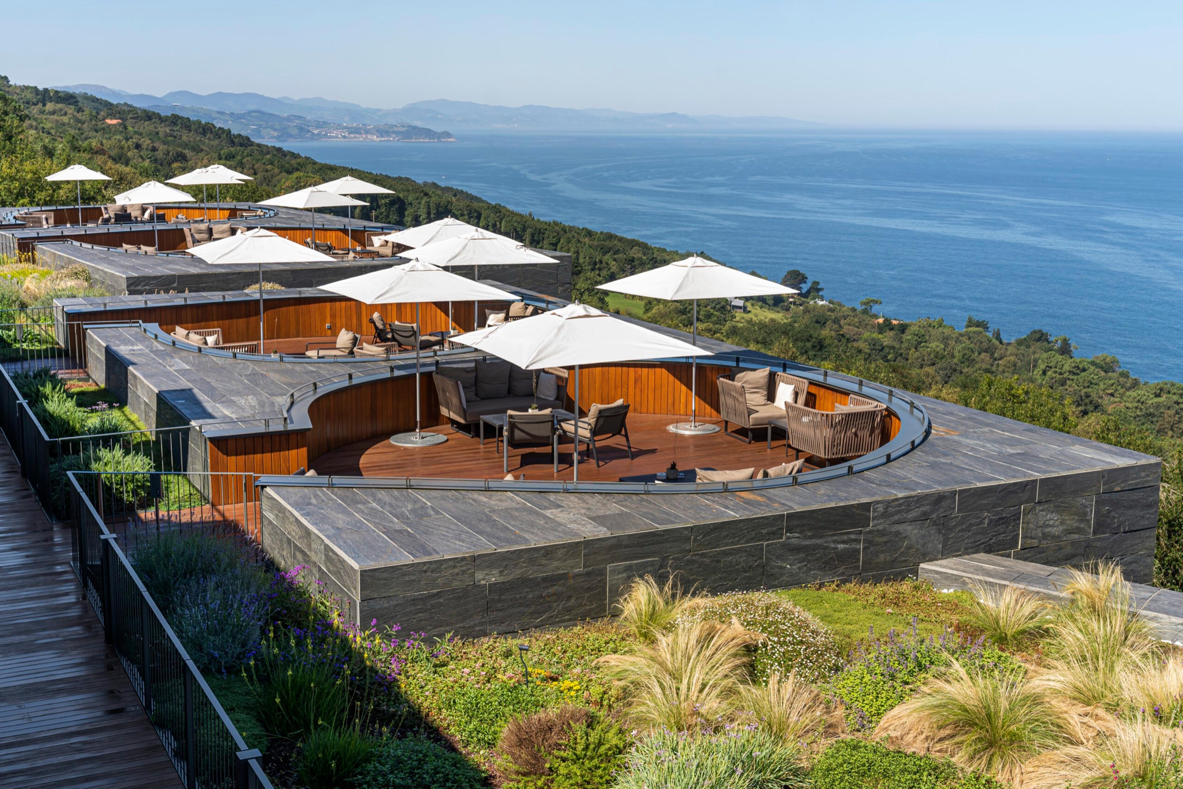 An aerial view of private terrace pods with sofas and umbrellas along a clifftop deck, overlooking the sea and a wooded coastline.