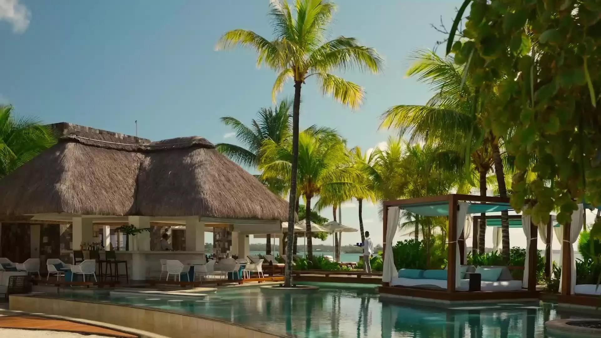 A thatched-roof bar and lounge beside a curved swimming pool, with cabana beds, palm trees and the sea beyond, and a waiter crossing the deck in the background.