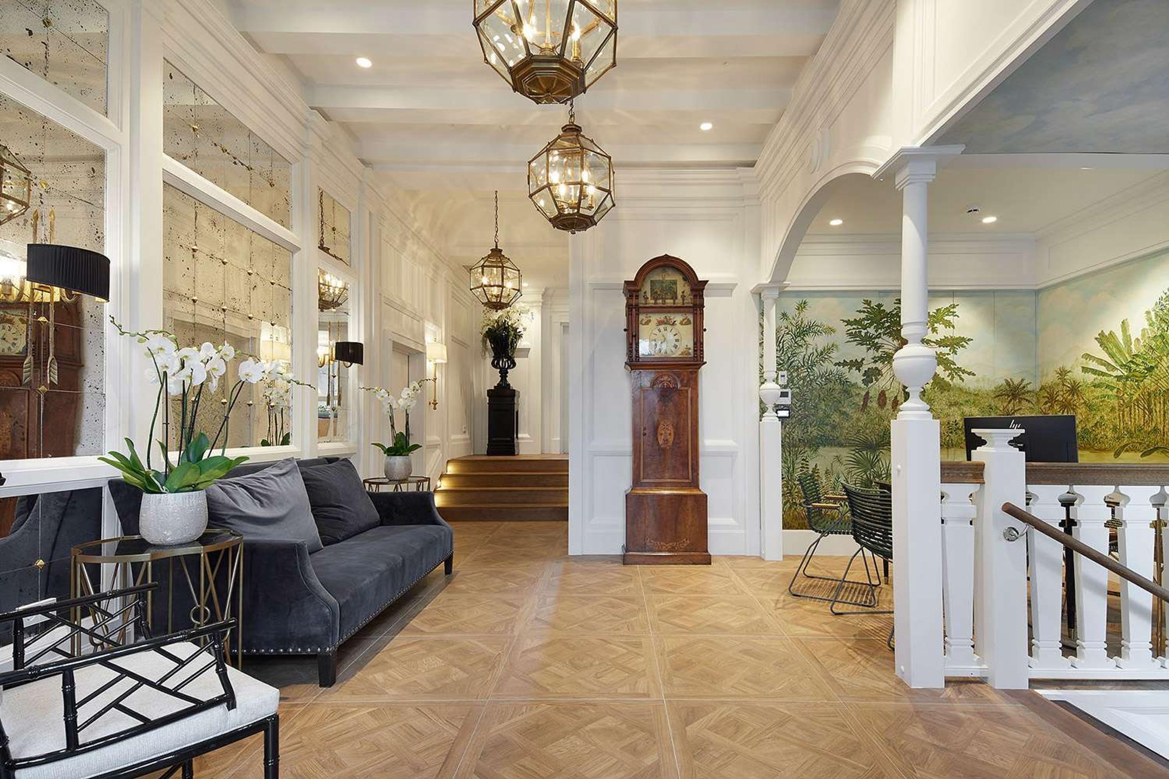 A hotel corridor with hanging lantern chandeliers, an antique grandfather clock, a mural of palm fronds, and steps up to a reception desk.