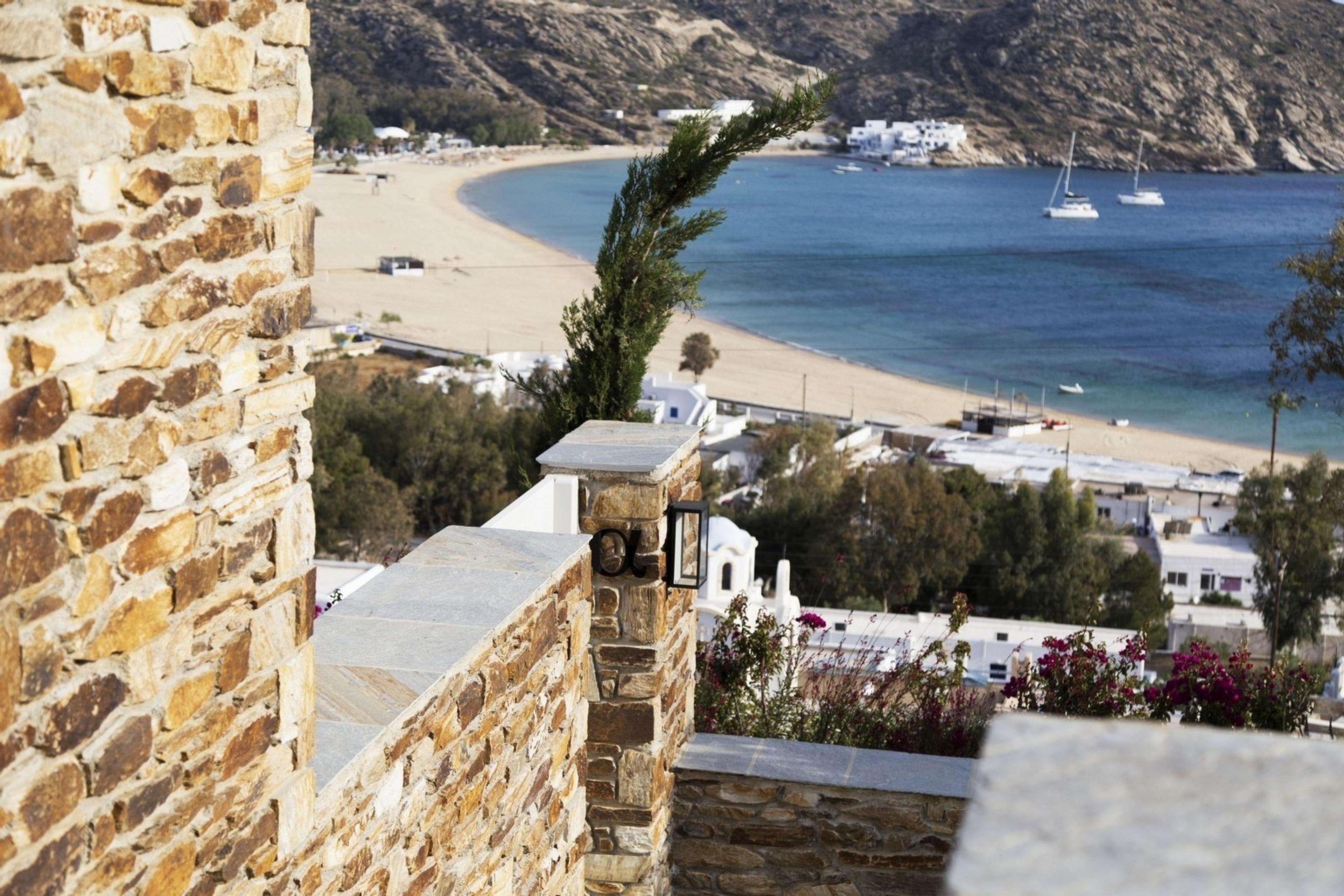 A view from a stone balcony down over a sandy bay, with boats on the water, a cypress tree and white houses along the shore.