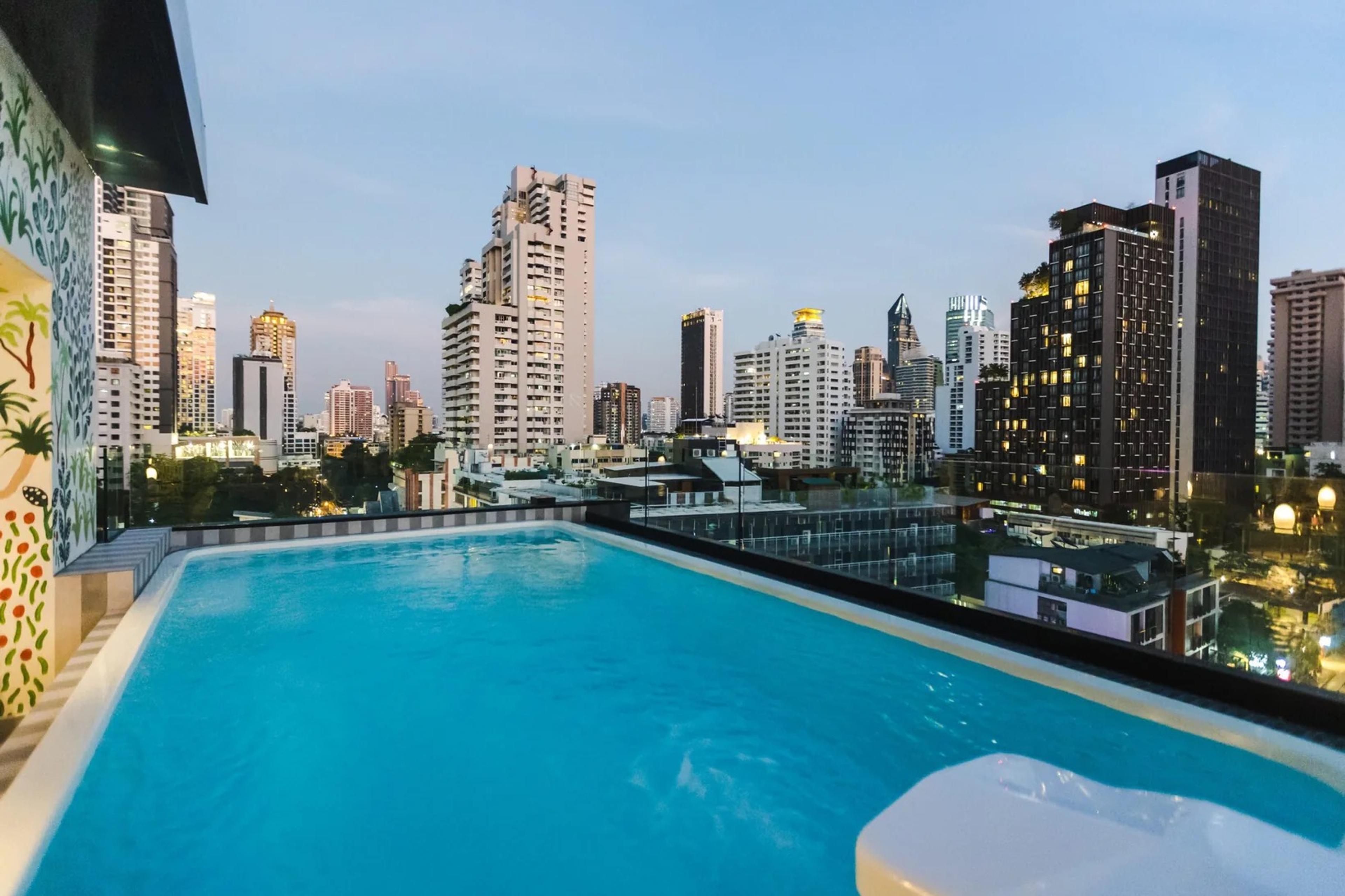 A rooftop swimming pool at dusk, with the lit towers of the Sukhumvit skyline rising behind it and a painted mural on the wall to the left.