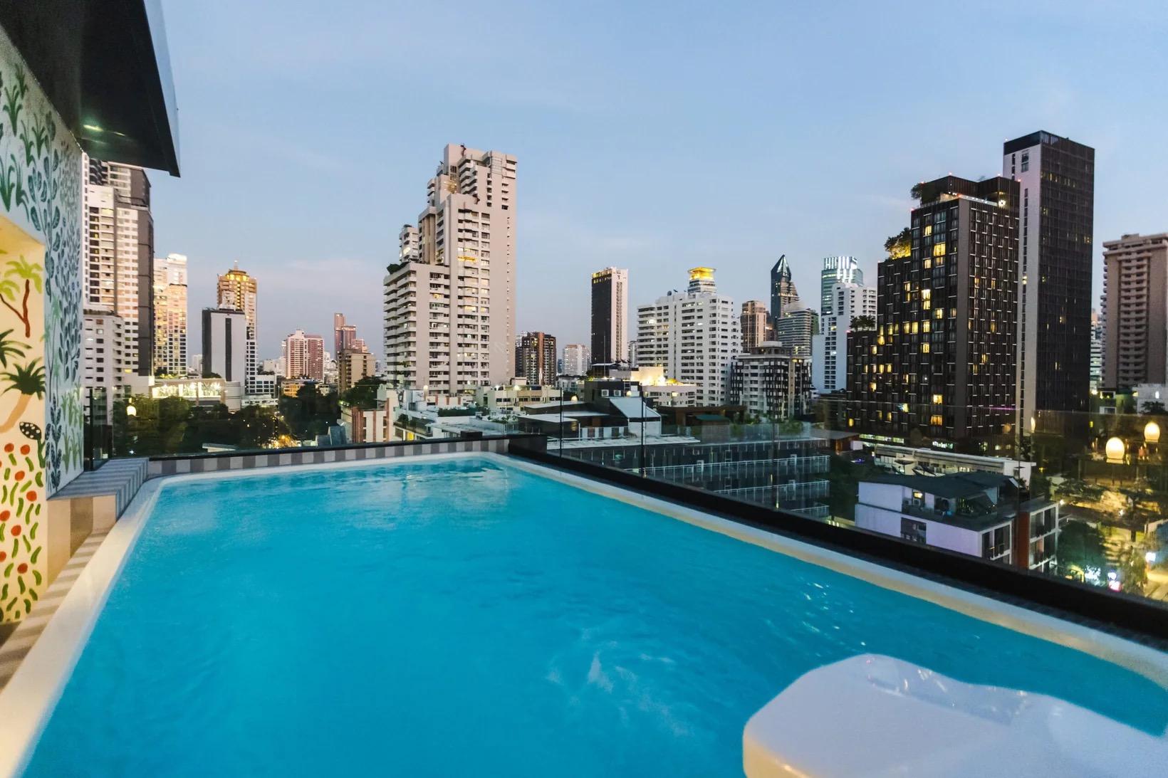 A rooftop swimming pool at dusk, with the lit towers of the Sukhumvit skyline rising behind it and a painted mural on the wall to the left.