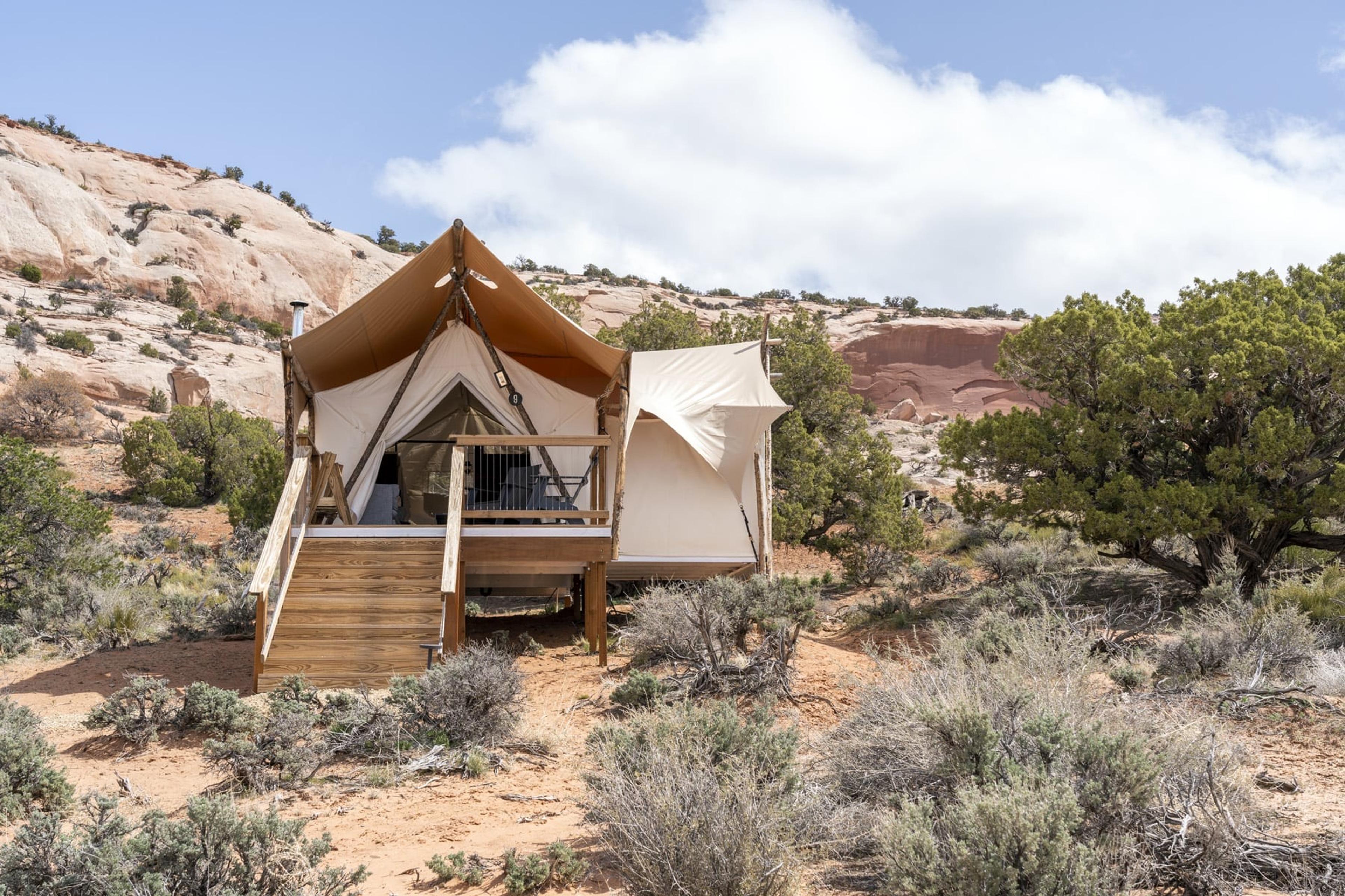 A canvas suite tent on a raised timber deck with wooden steps, set among juniper and scrub at the foot of pale sandstone cliffs under a cloudy sky.