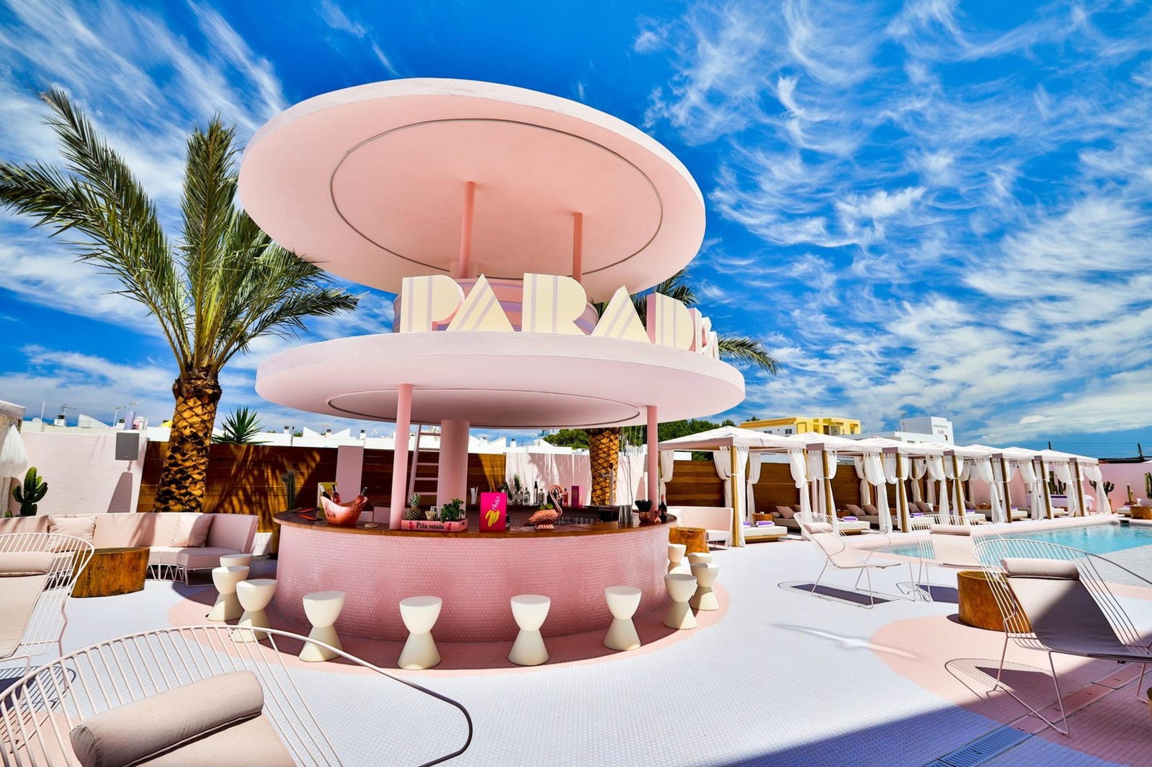 A round pink bar under a two-tier pink roof with striped lettering, with white stools around it, on a white terrace; a pool and white cabanas behind, a palm on the left and blue sky above.