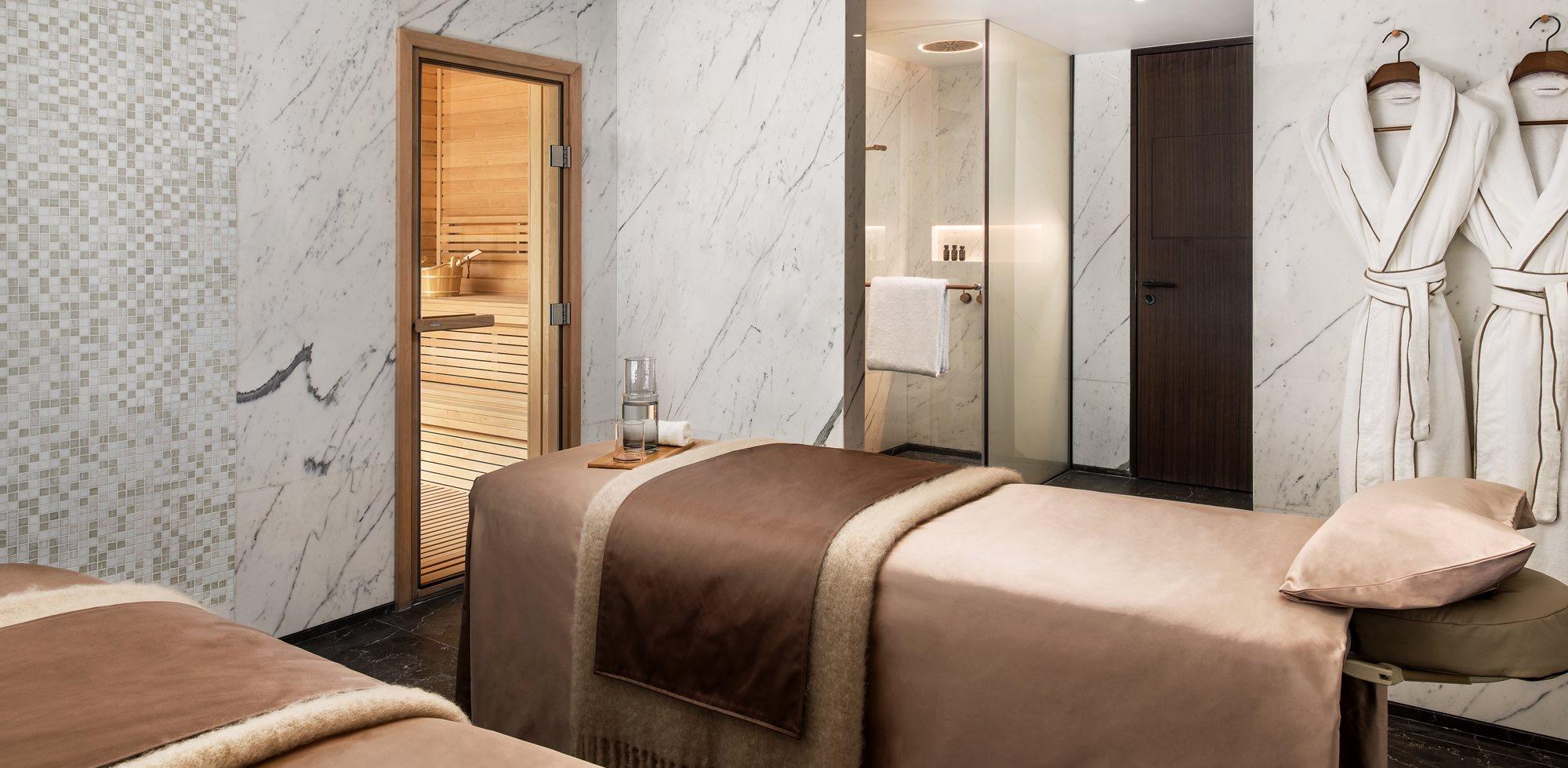 a spa treatment room with two massage beds, white robes hanging by a sauna door, and marble walls
