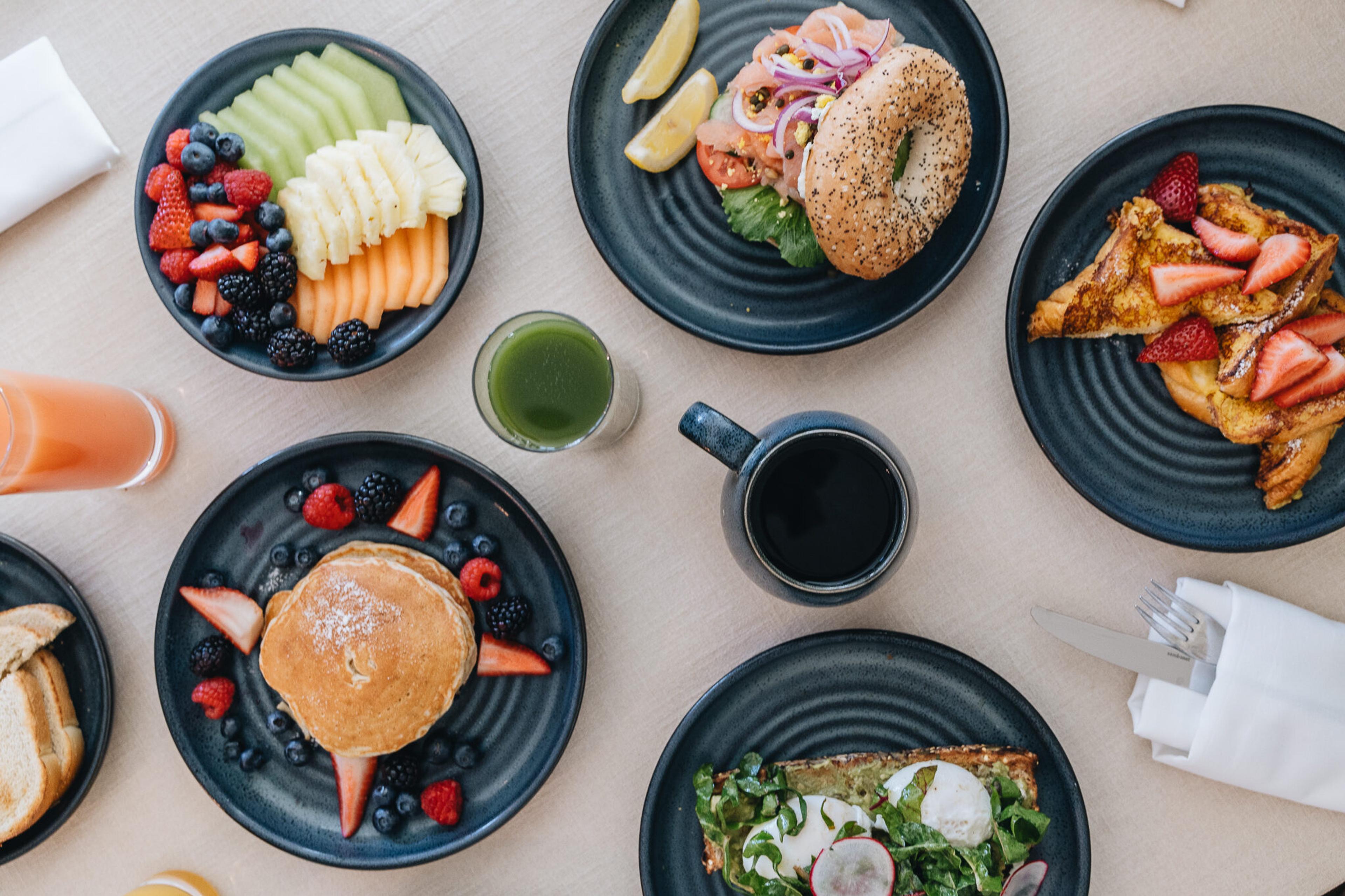 Overhead view of breakfast on a pale table: pancakes with berries, a bagel with smoked salmon and red onion, French toast with strawberries, a bowl of fruit, a green juice and a black coffee.
