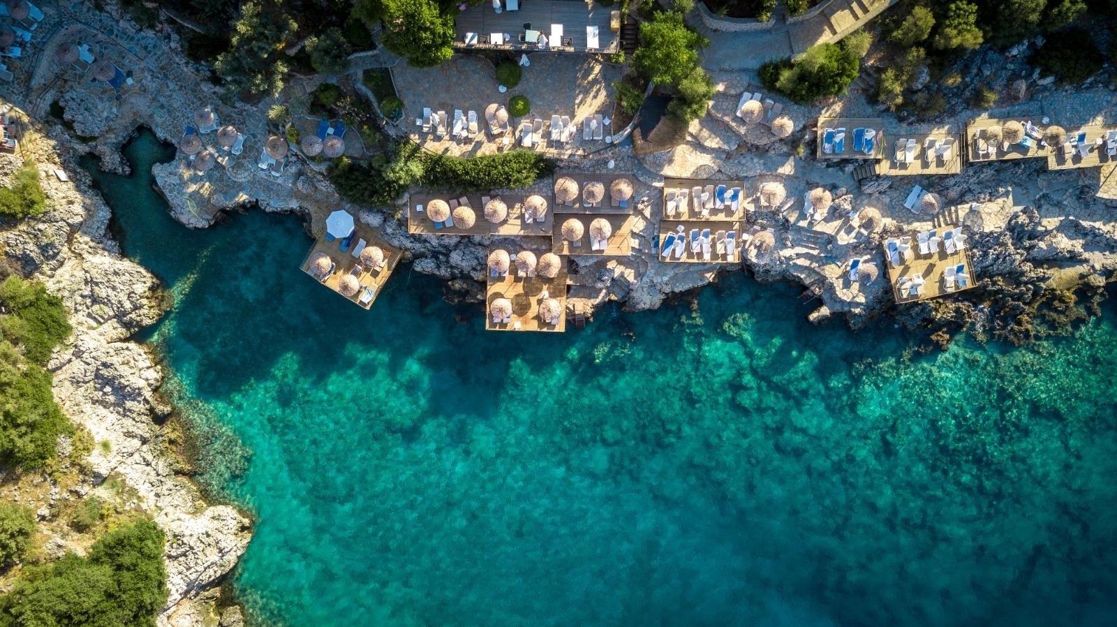 An aerial view of wooden sunbathing platforms and umbrellas built into a rocky shoreline above clear turquoise water.