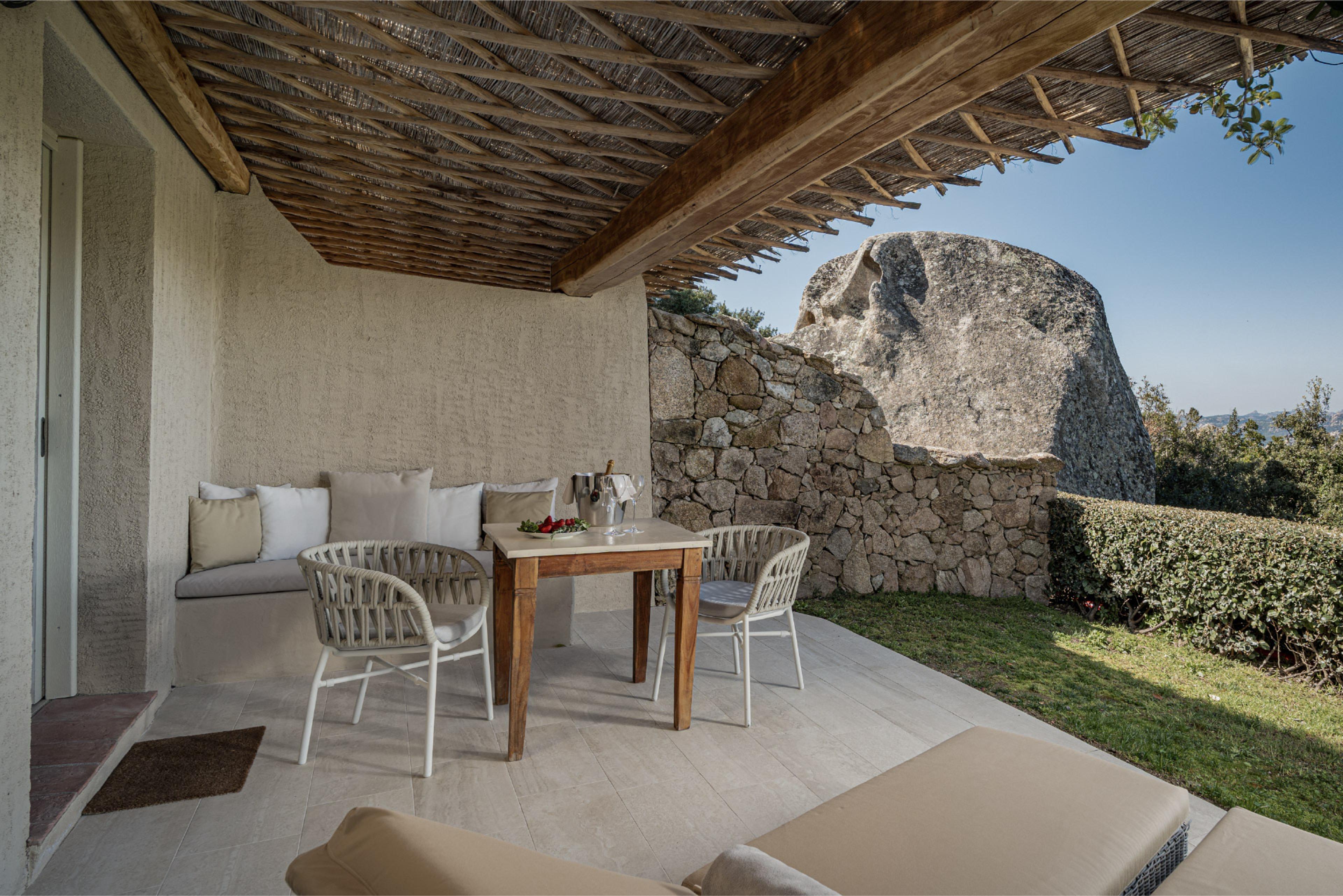 A covered terrace with a dining table and chairs, a built-in stone bench, and a large granite boulder in the garden beyond.