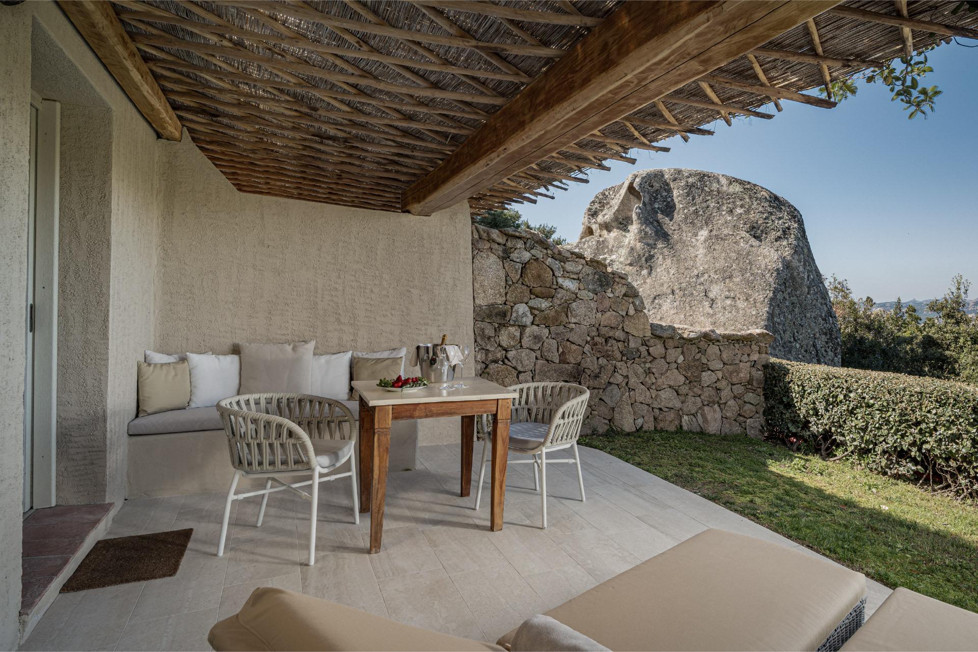 A covered terrace with a dining table and chairs, a built-in stone bench, and a large granite boulder in the garden beyond.
