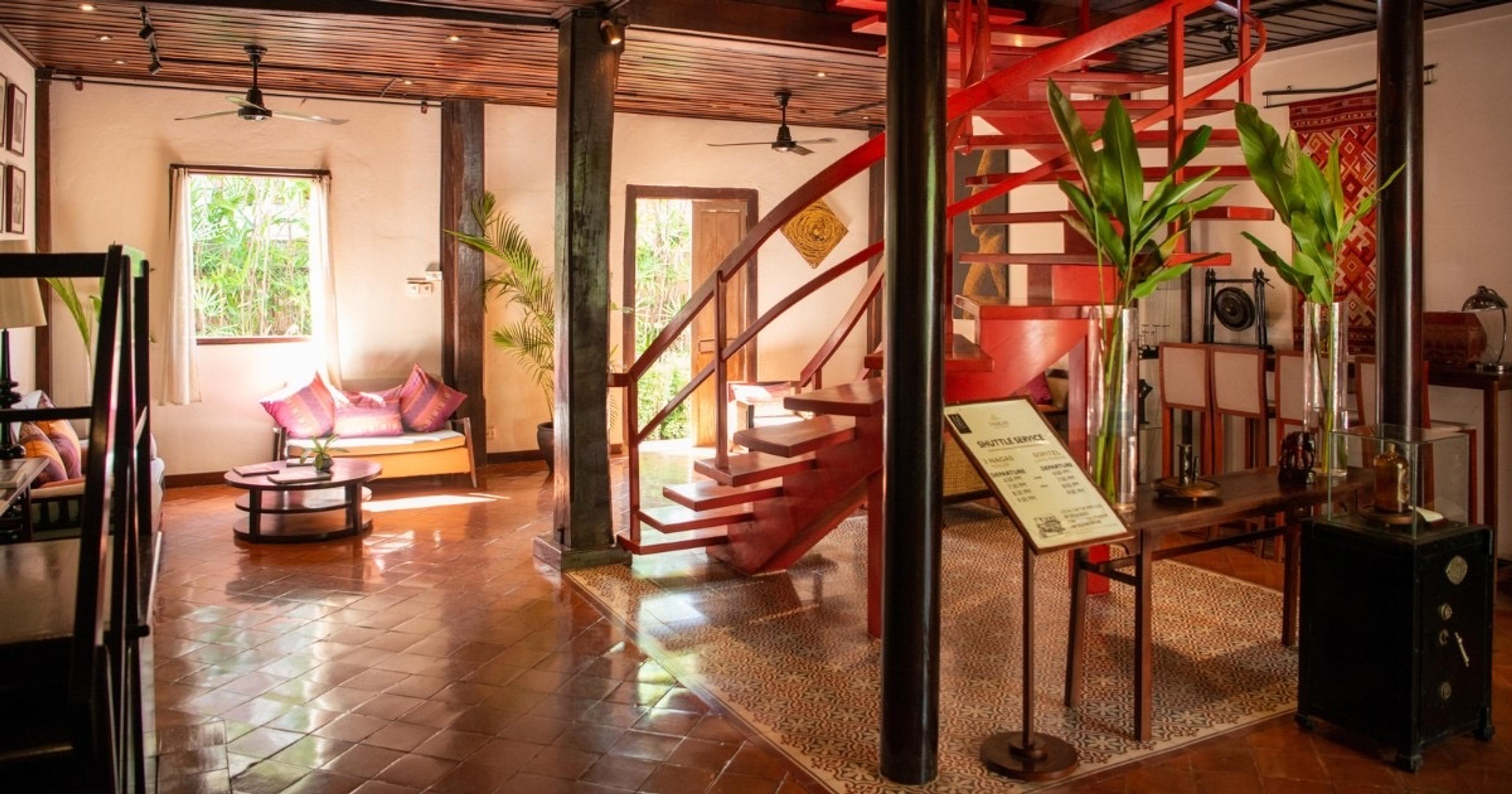 A hotel lobby with a red spiral staircase, dark wood columns, patterned floor tiles, and a sofa with pink cushions by a window.