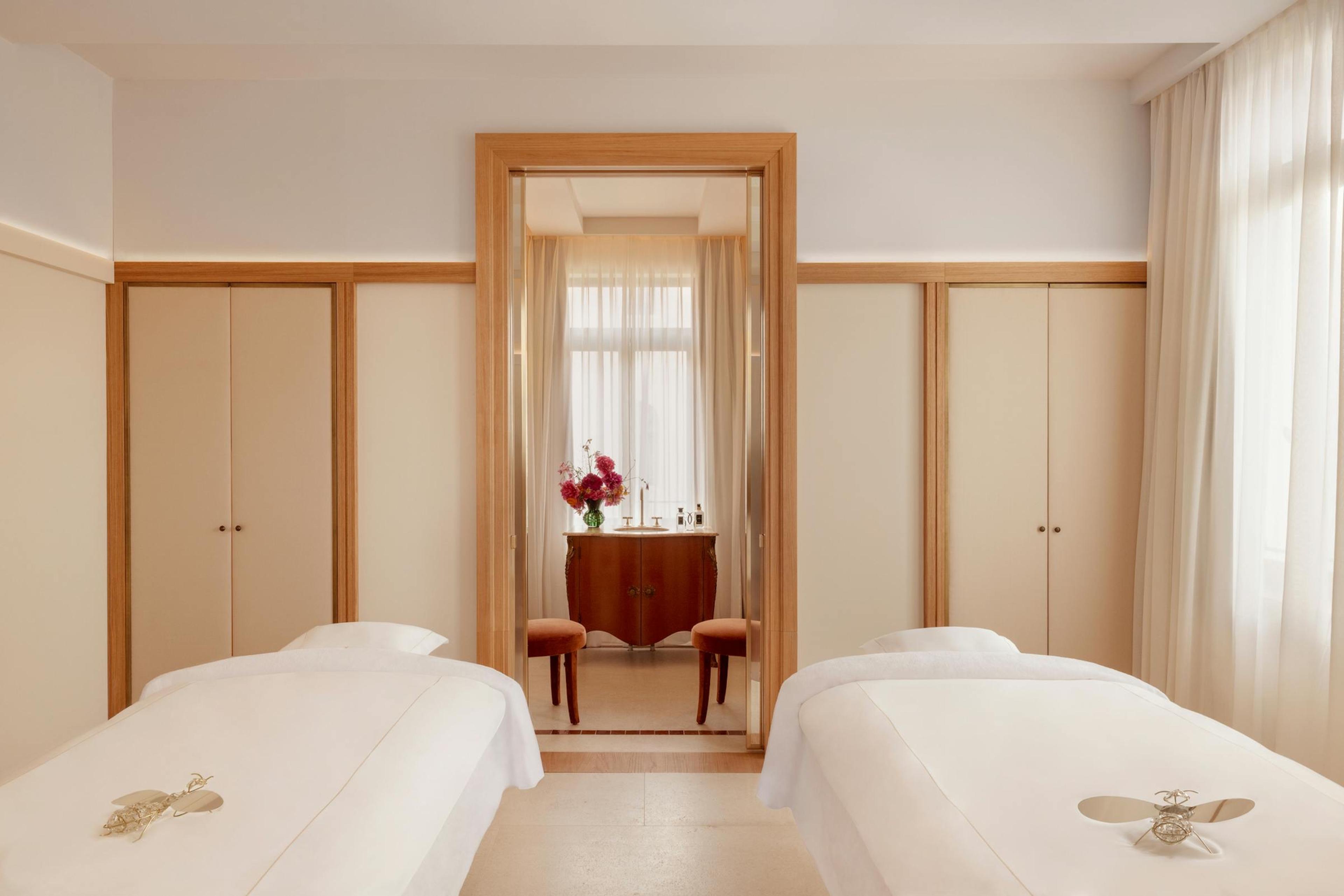 A spa room with two massage beds dressed in white linen on a terrazzo floor, and a doorway through to a washstand with flowers.