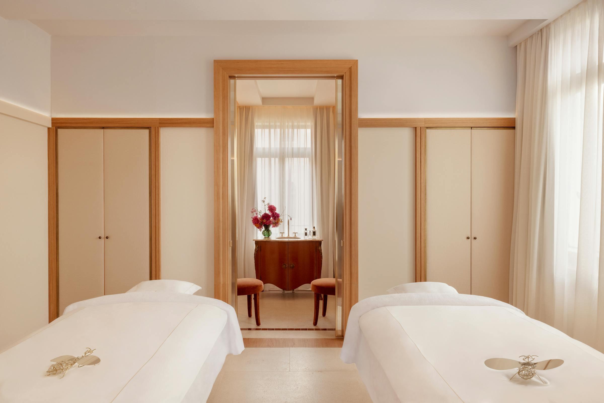 A spa room with two massage beds dressed in white linen on a terrazzo floor, and a doorway through to a washstand with flowers.