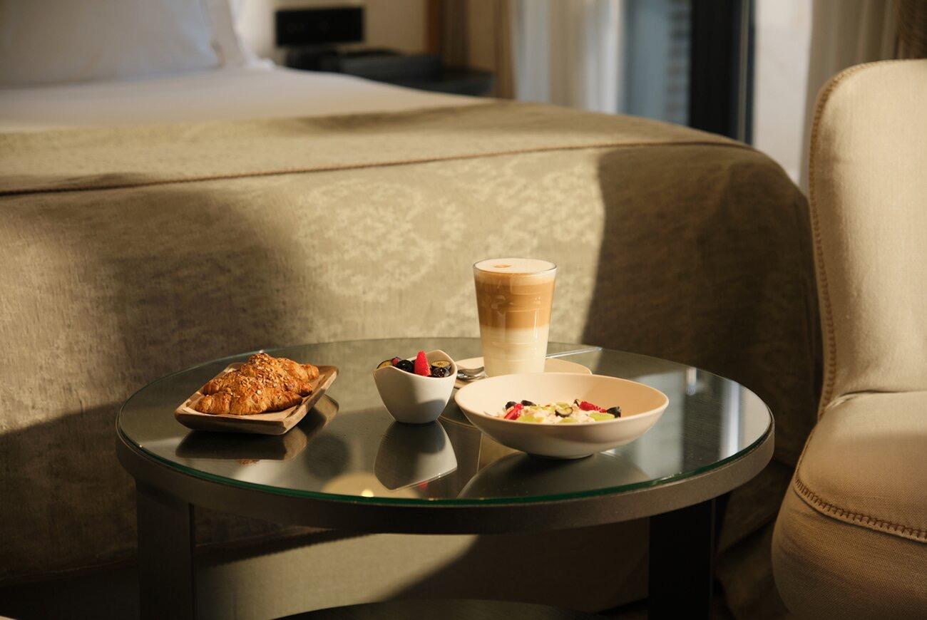 A glass-topped table beside a bed holds a basket of pastries, a bowl of berries, a bowl of muesli topped with fruit, and a tall glass of layered coffee, with a cream armchair alongside.