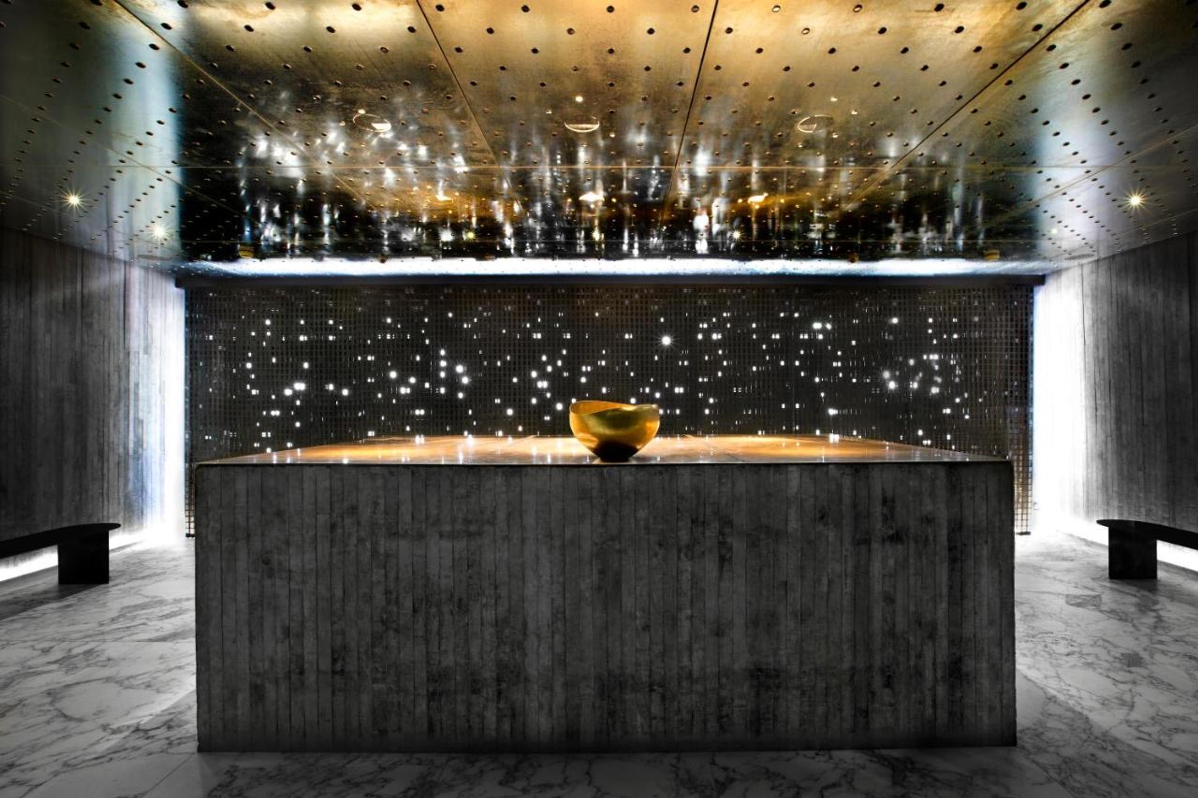 A concrete reception counter holding a round brass bowl, lit beneath a gold dotted ceiling and a wall of pinpoint lights, with marble flooring and benches either side.