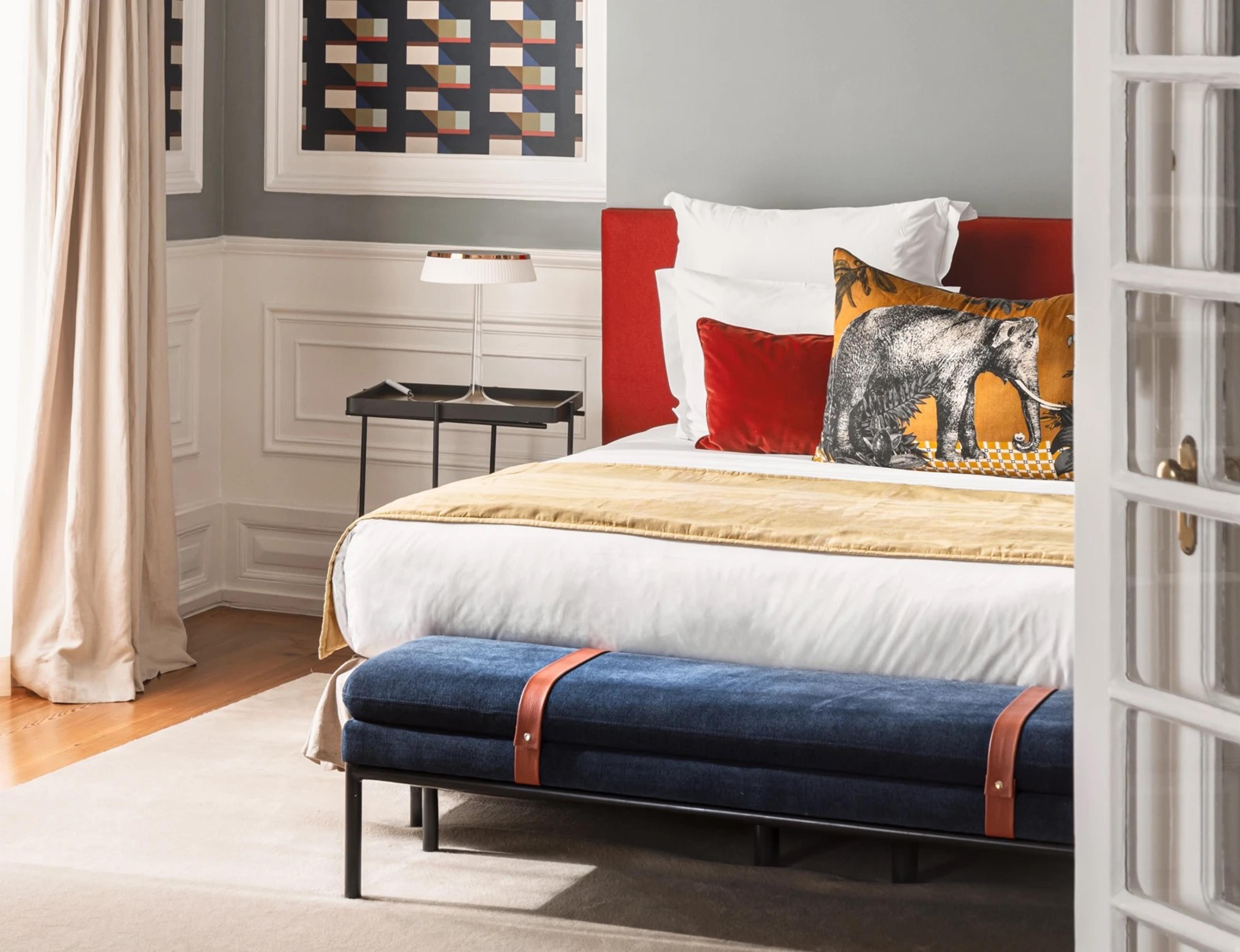 A bed with a red headboard, a gold throw, and a cushion printed with an elephant, beside a navy bench with tan leather straps and a pale wall panelled in white.
