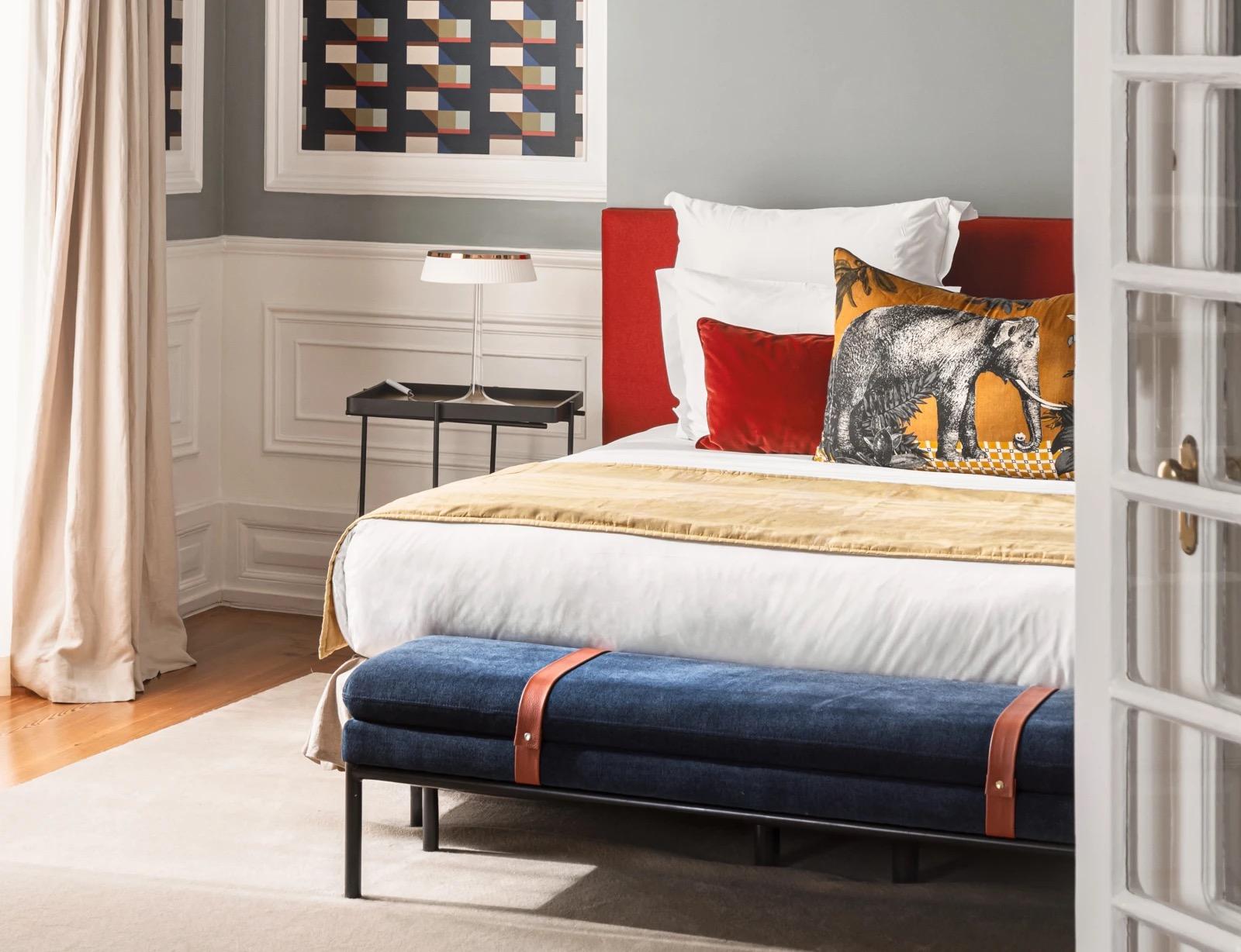 A bed with a red headboard, a gold throw, and a cushion printed with an elephant, beside a navy bench with tan leather straps and a pale wall panelled in white.
