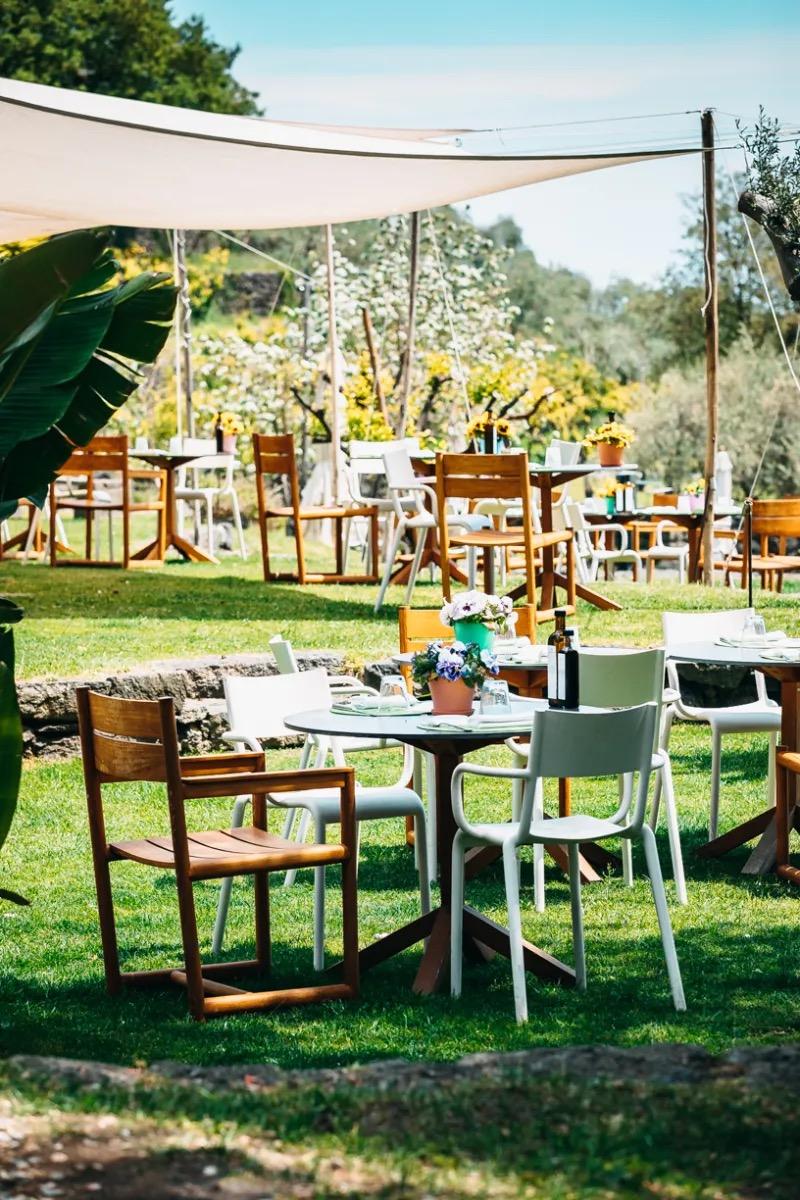 A shaded outdoor dining area with wooden and white chairs around round tables set for lunch on a lawn.