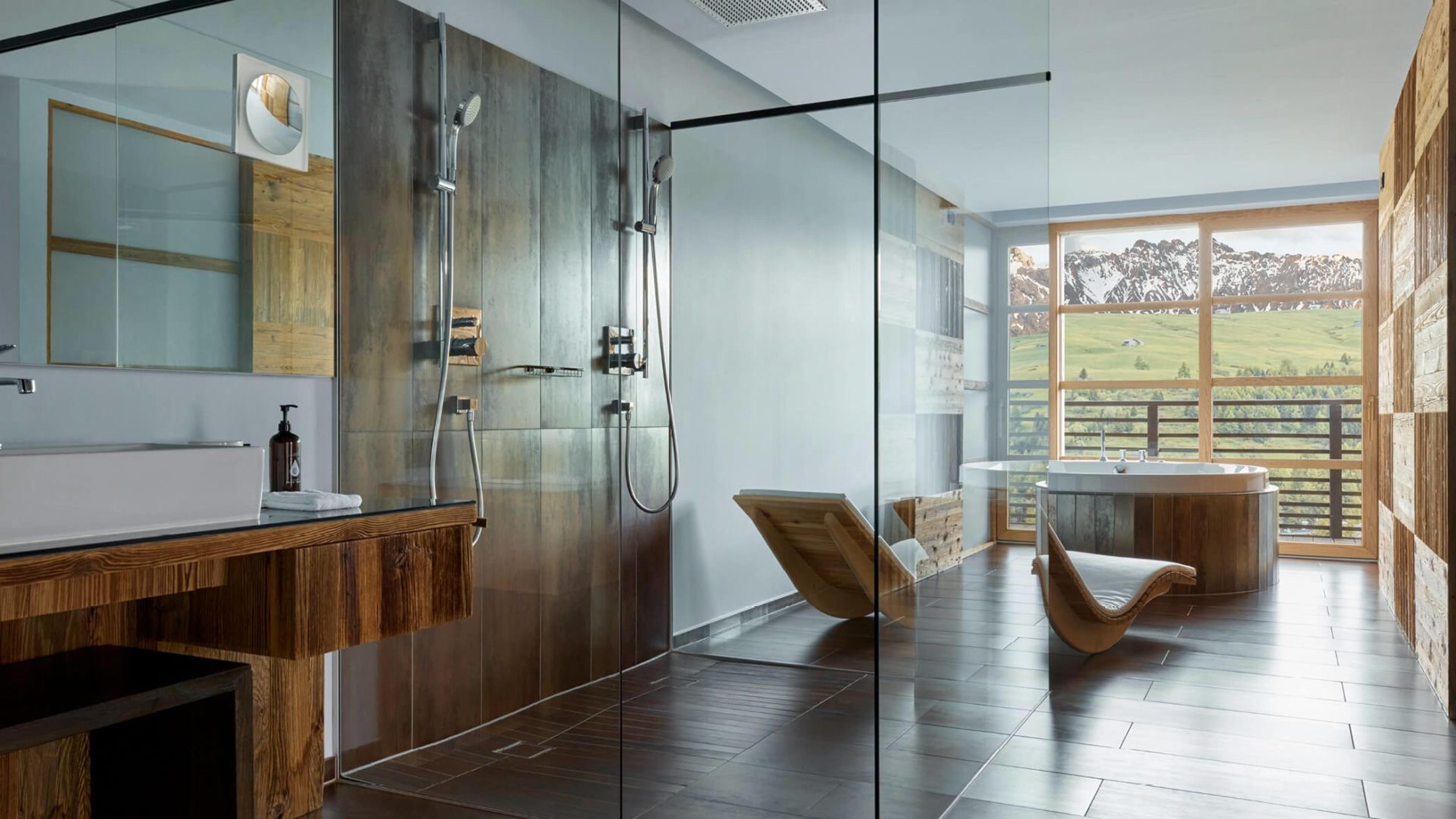 A round wood-clad freestanding tub and two wooden lounge chairs sit before a large window onto snow-covered mountains, beside a tiled walk-in shower.