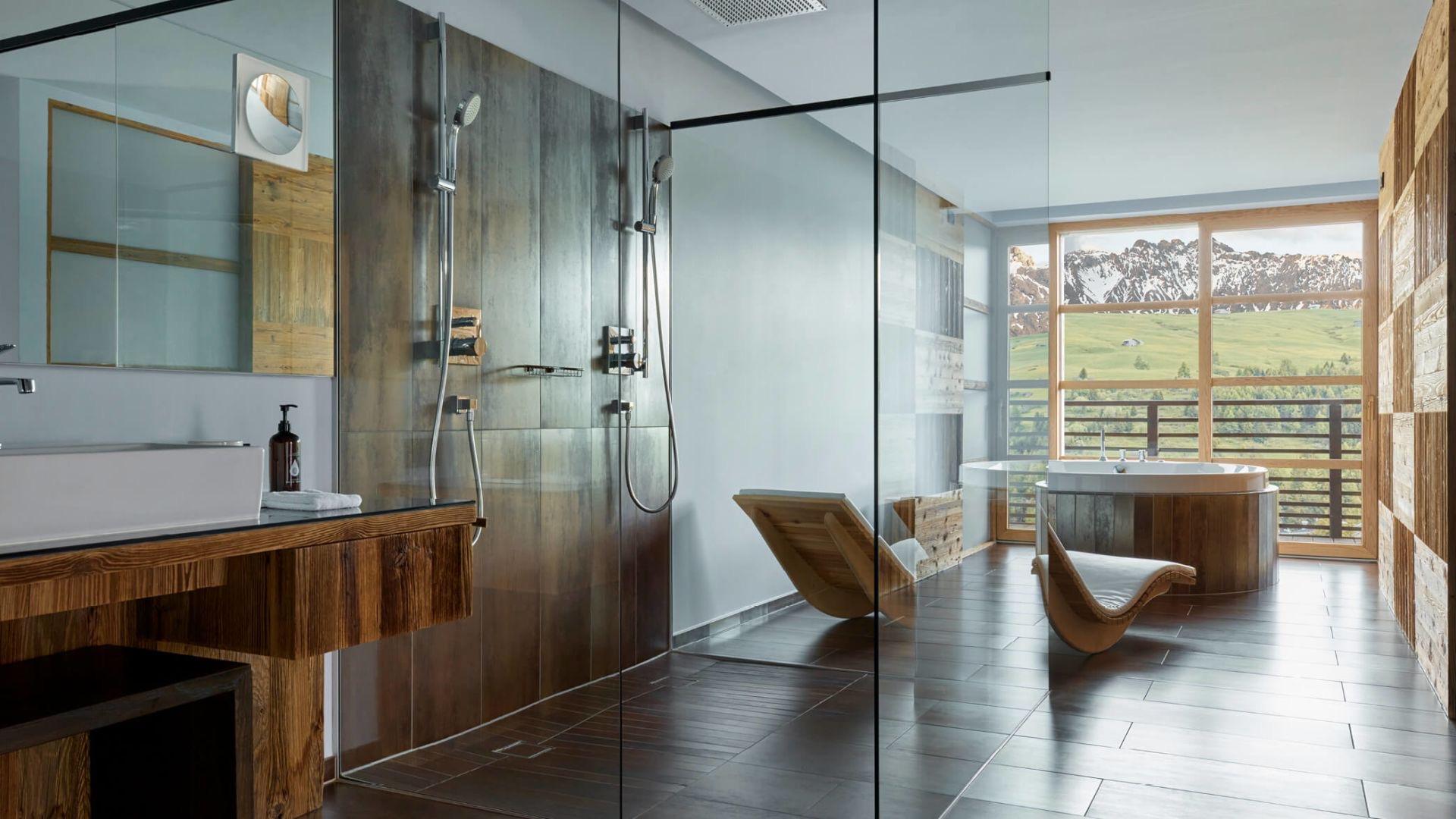A round wood-clad freestanding tub and two wooden lounge chairs sit before a large window onto snow-covered mountains, beside a tiled walk-in shower.