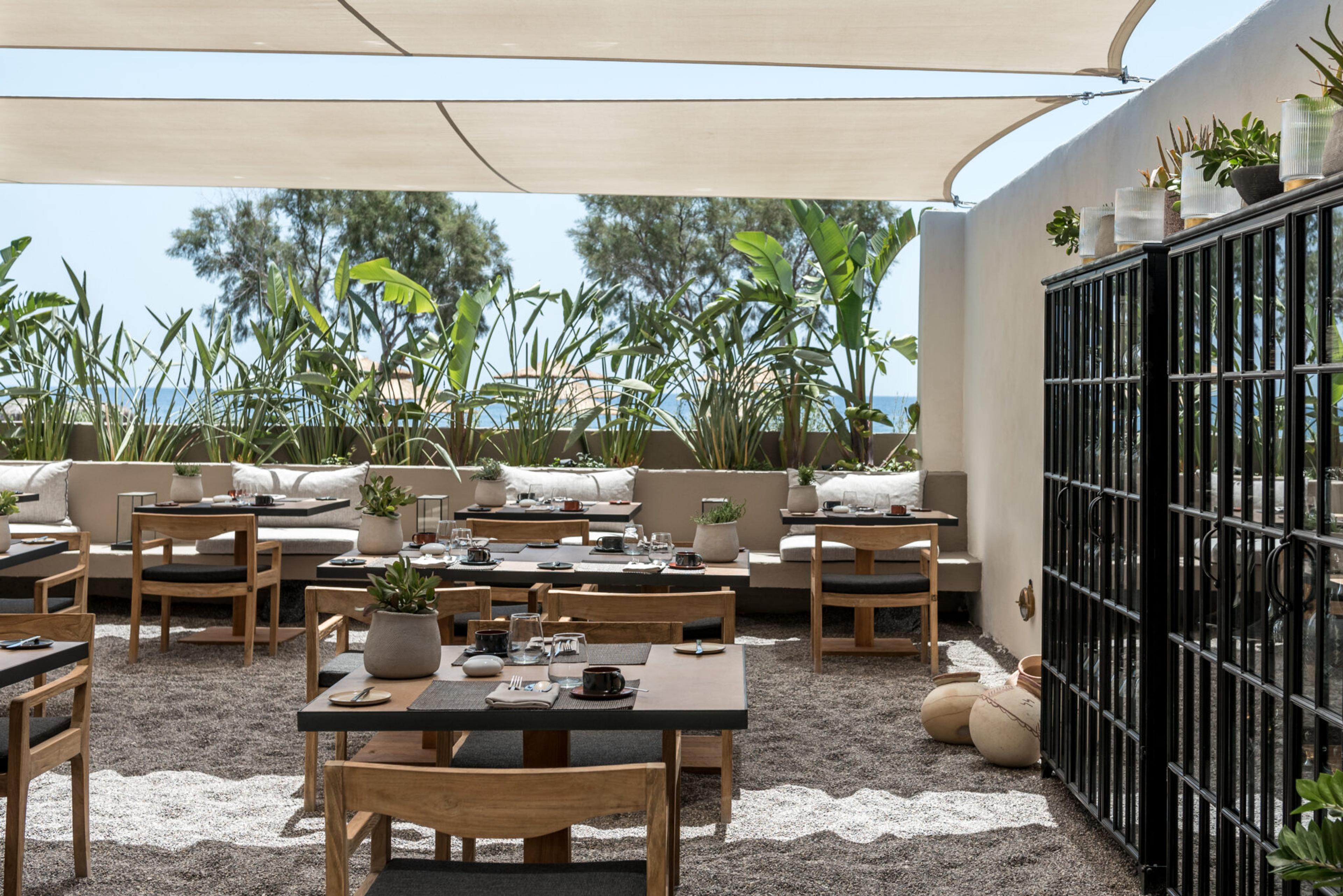 An outdoor dining terrace with wooden tables set for a meal, a sail-cloth canopy overhead and tropical plants behind a low wall.