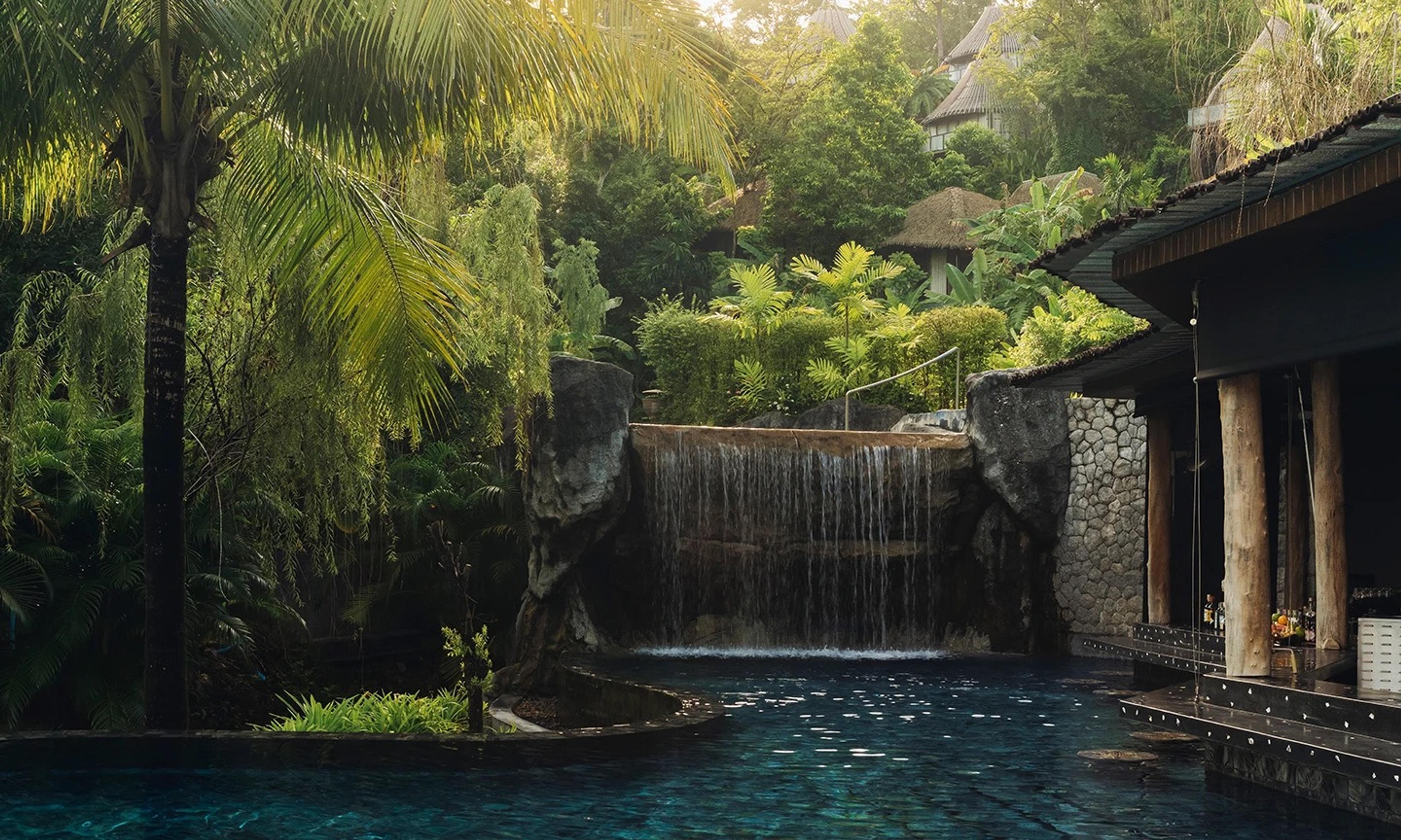 A rock waterfall feeding an outdoor pool beside a thatched poolside bar, framed by palm trees.