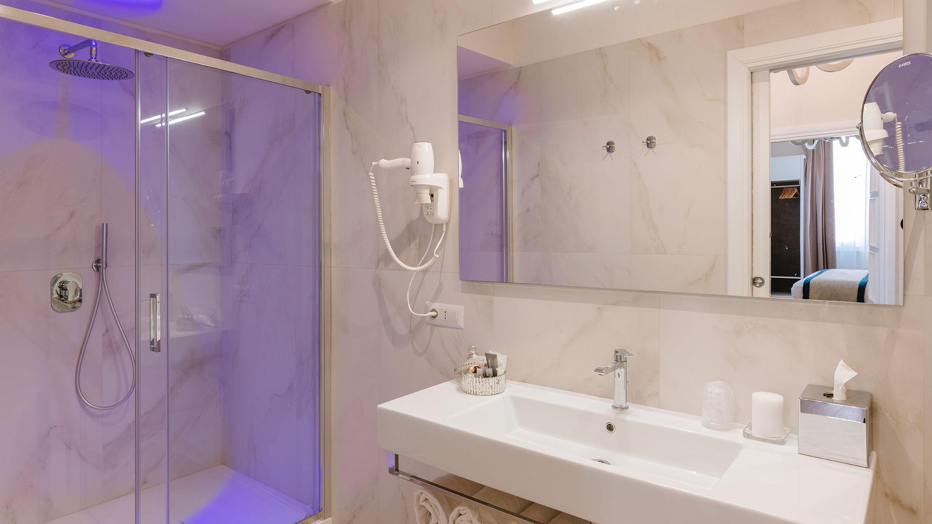 A bathroom with a glass-walled shower lit blue, a long marble-topped sink, and a wide mirror reflecting the bedroom beyond.