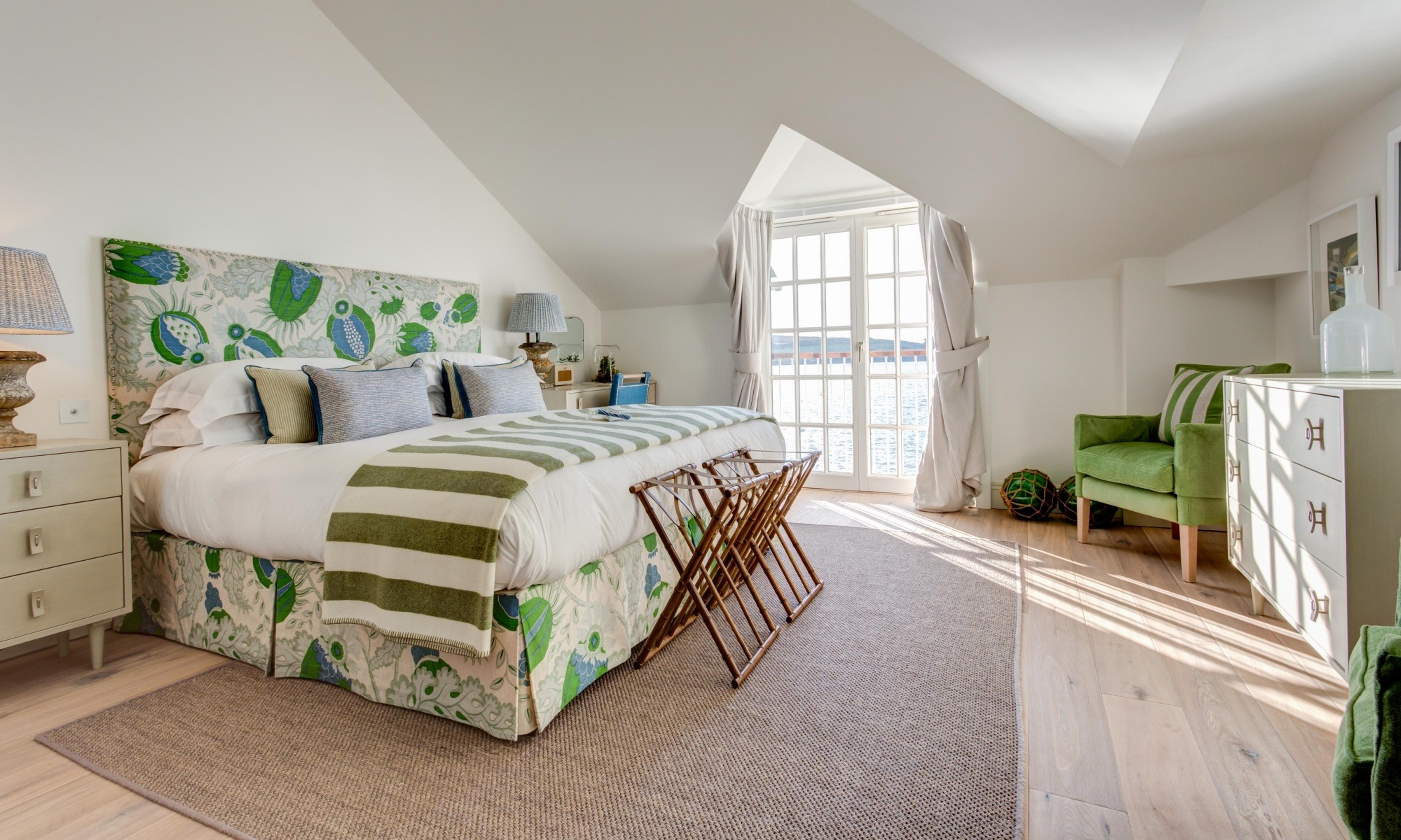 A bedroom under a sloped ceiling with a green and blue floral headboard, a striped throw on the bed, a green armchair, and open French doors leading to a balcony.