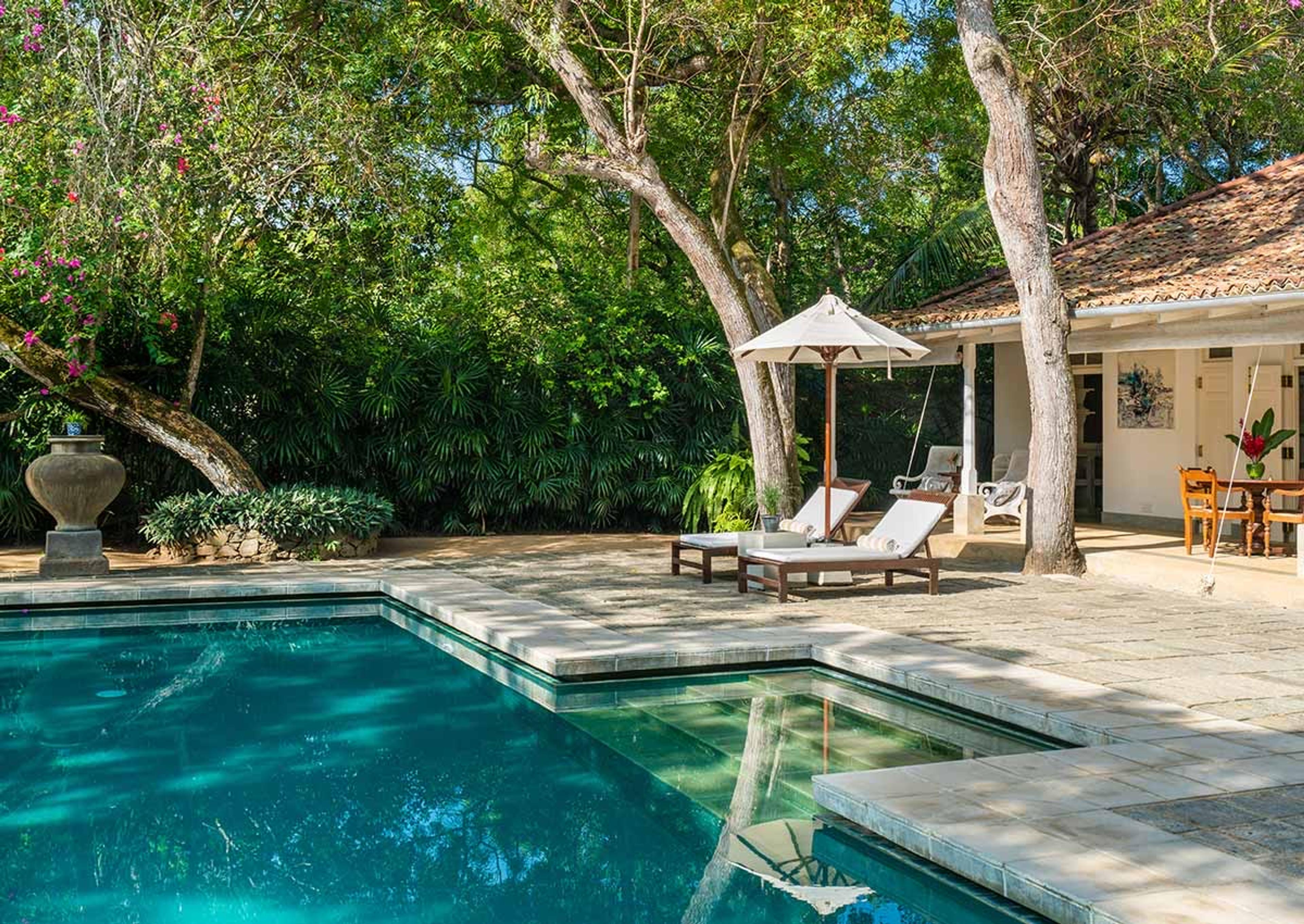 In daylight, a pool with a stone urn at its end, bougainvillea on the left, and a tiled-roof pavilion with loungers and a parasol on the right.