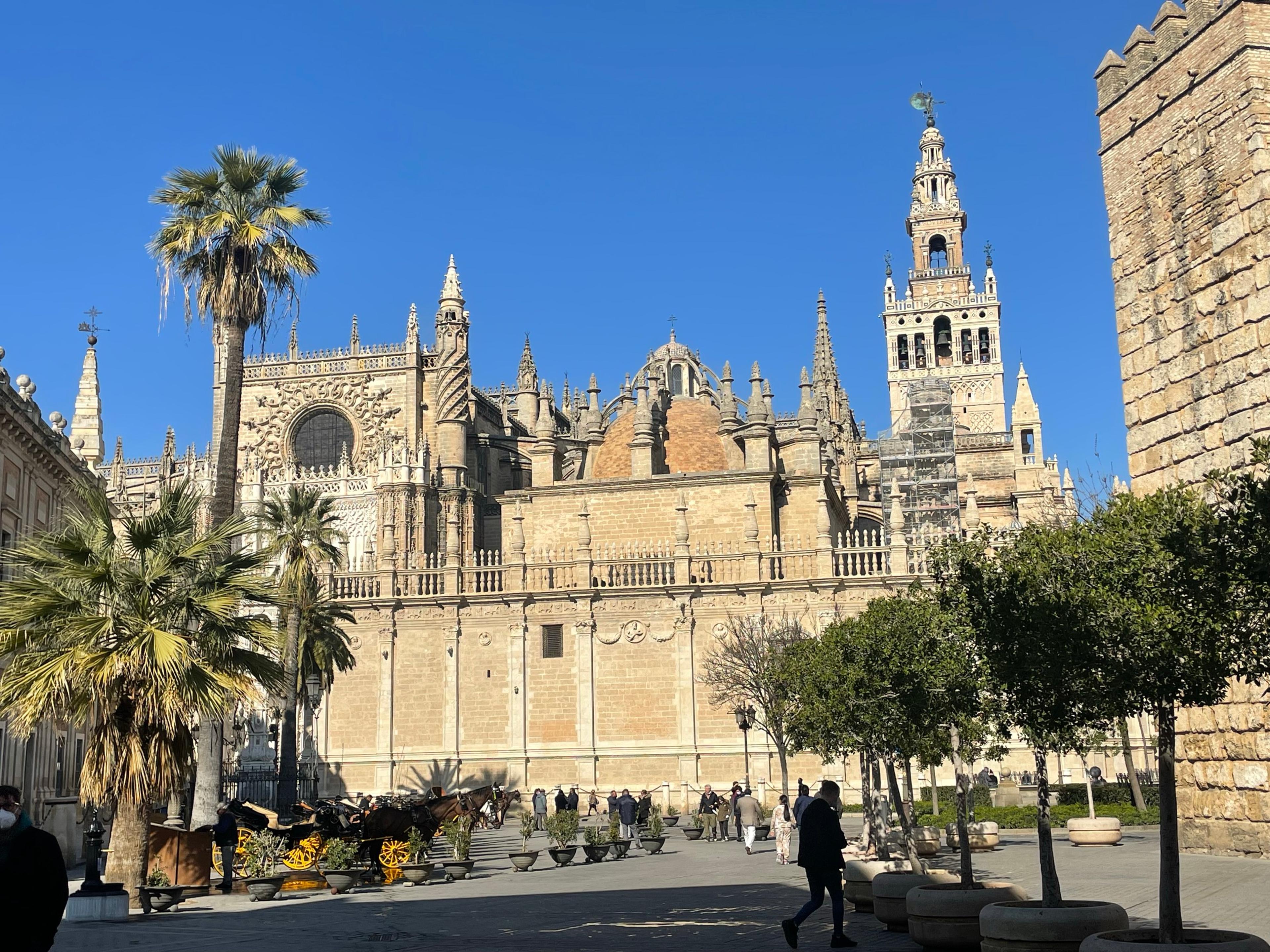 Seville Cathedral and the Giralda from the square beside the Alcázar walls, with palms and horse carriages