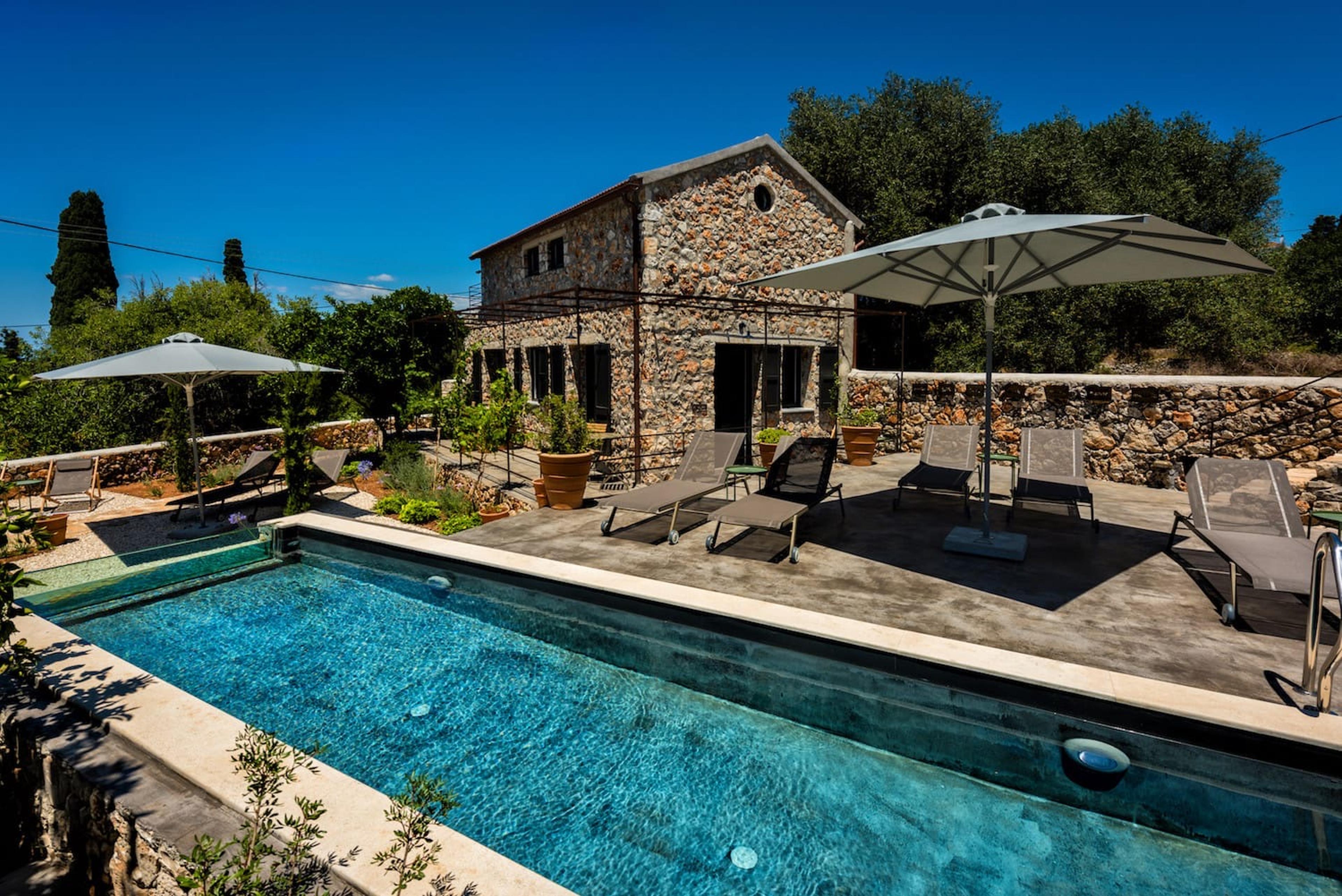 A two-storey stone house with a round window in its gable end, seen across a terrace with sun loungers, umbrellas and a long pool in full sun.