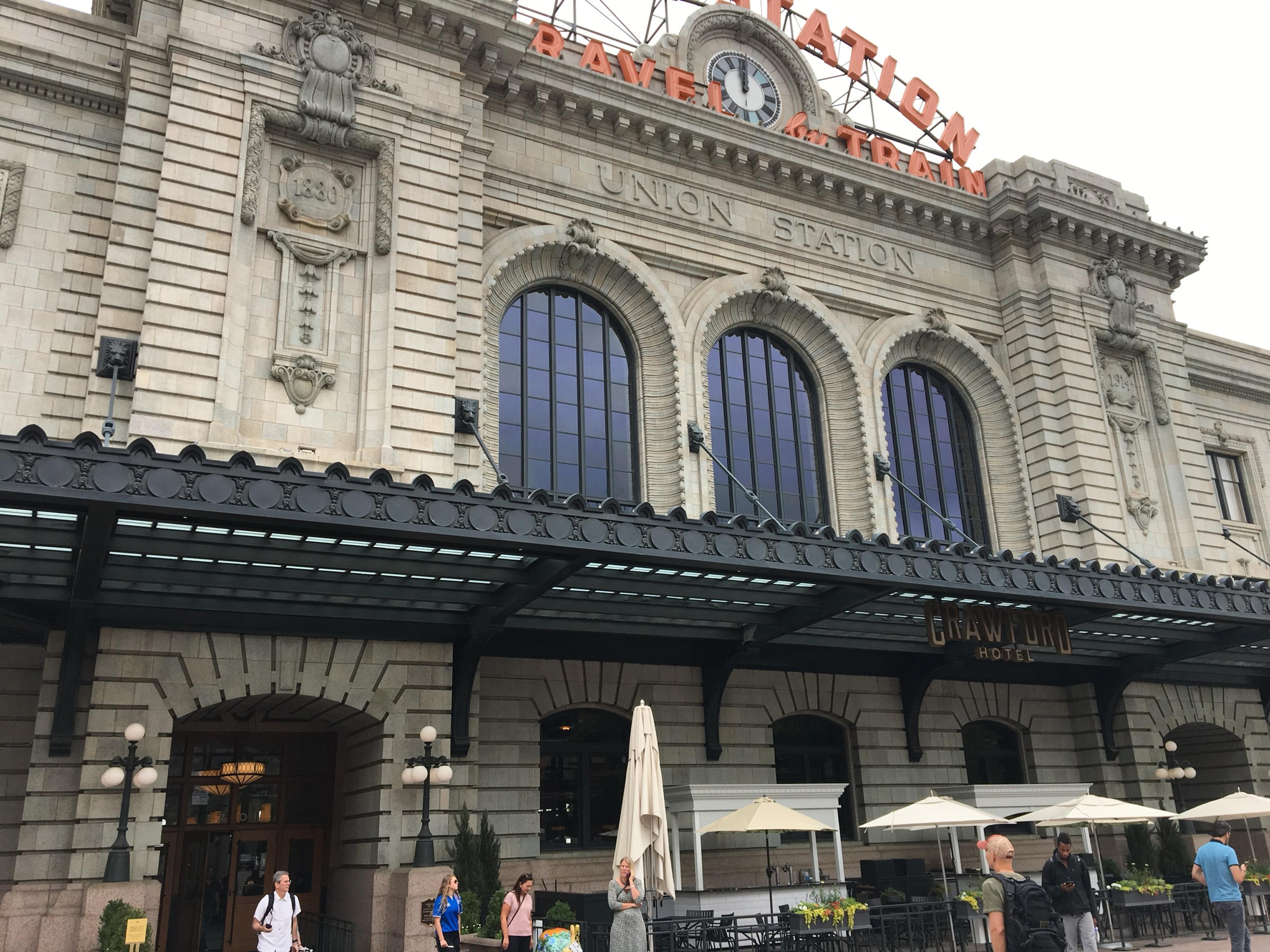 The front of Denver's Union Station, with its Travel by Train sign and clock above the arched windows and café umbrellas below